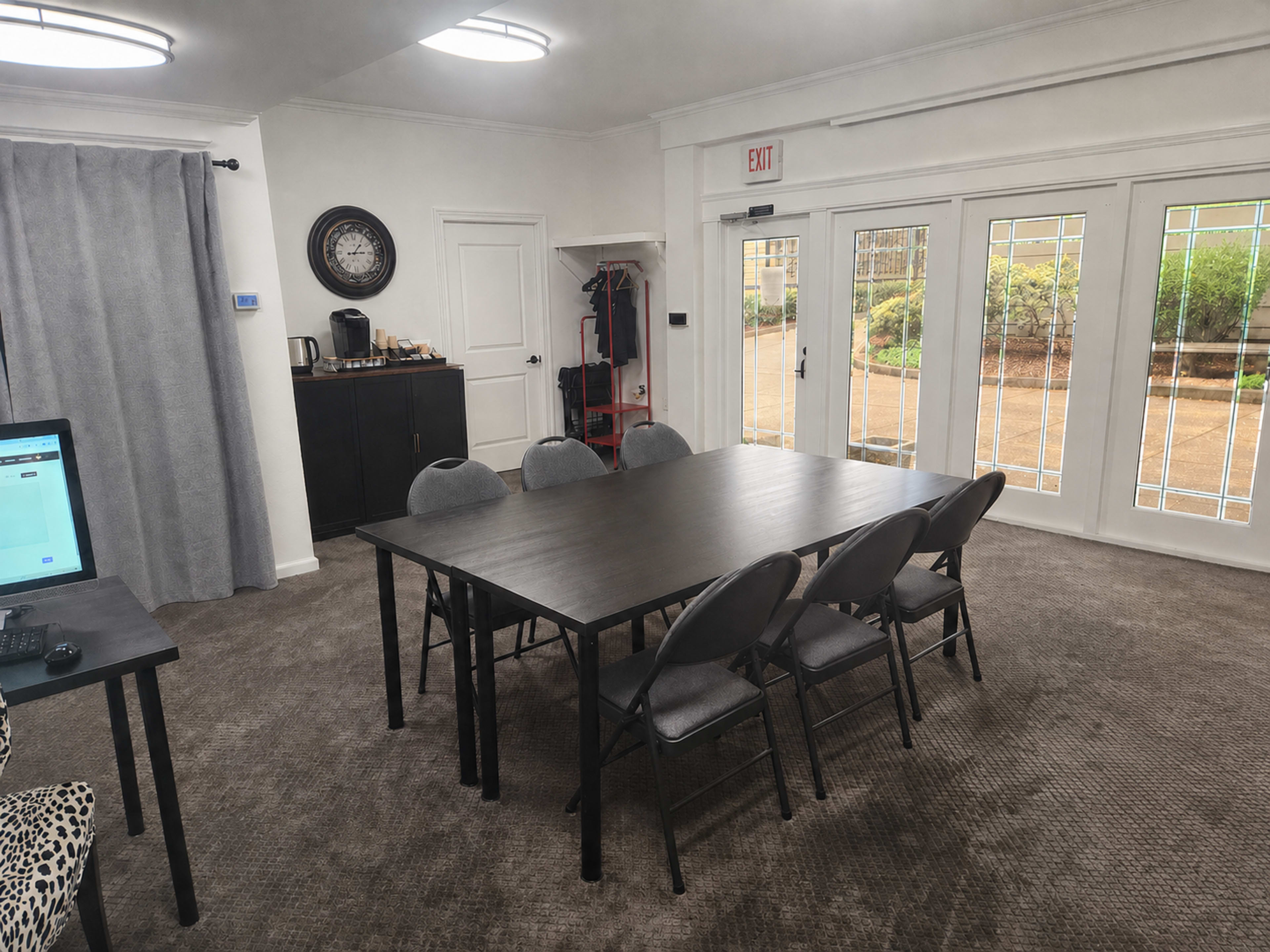 The image shows a meeting room with a large rectangular table surrounded by grey chairs, a sideboard, and glass double doors leading to an outdoor area.
