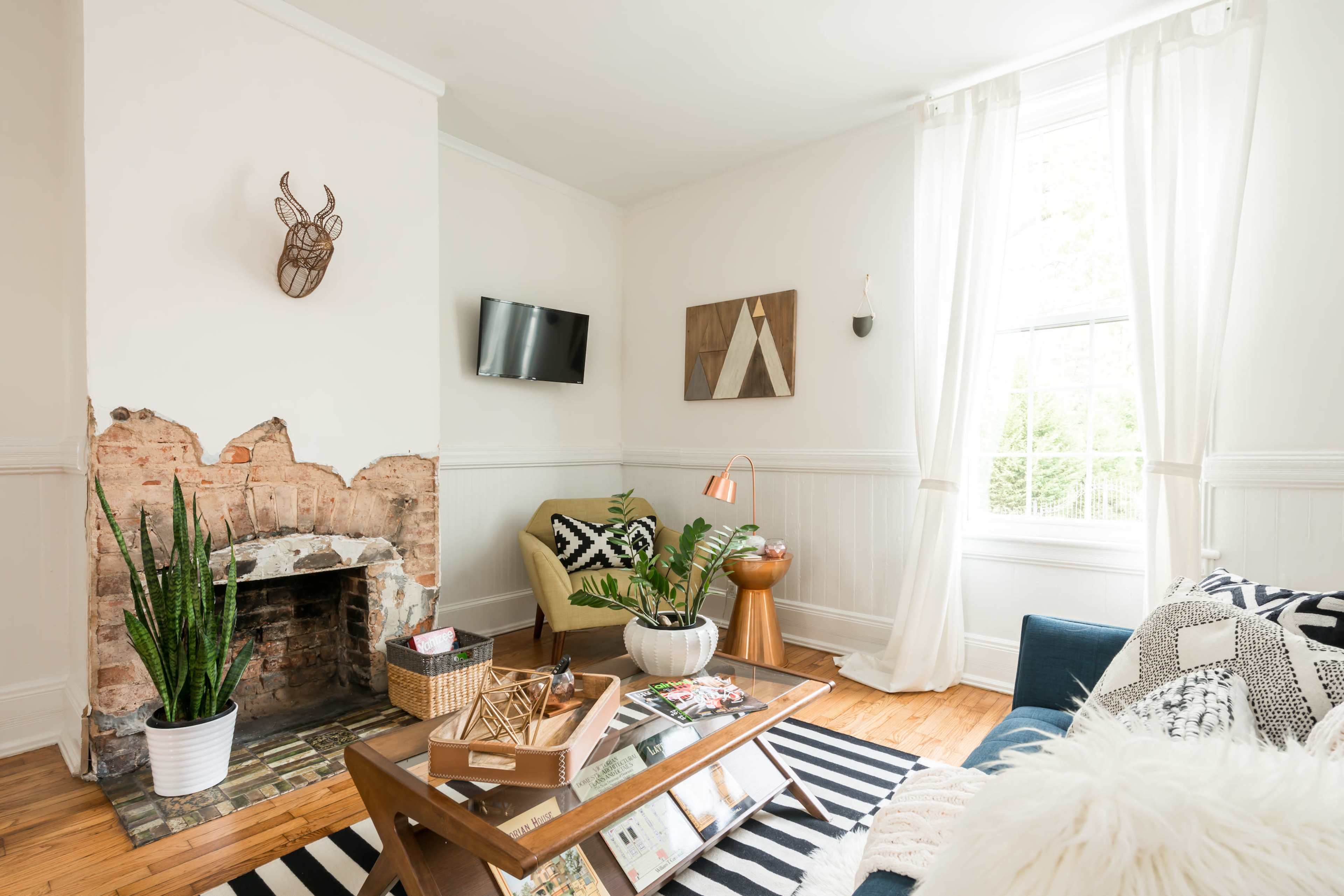 The image shows a bright living room featuring a mounted decorative mask, a small flat-screen TV, a fireplace with exposed brick, and various furniture pieces, including a green armchair and a coffee table with books and magazines.