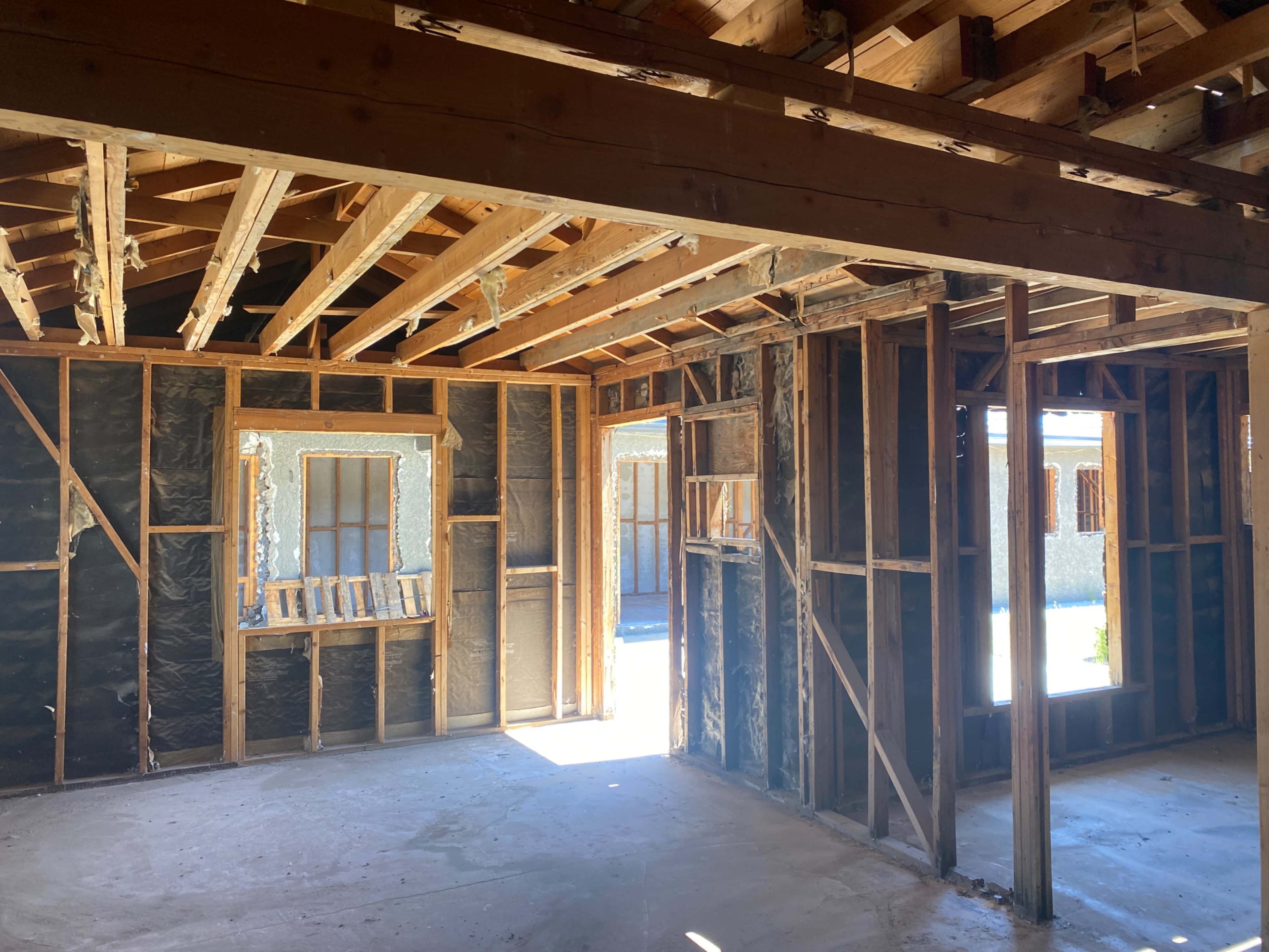 The interior of a house under renovation shows exposed wooden framing and insulation, with openings leading to outside areas.
