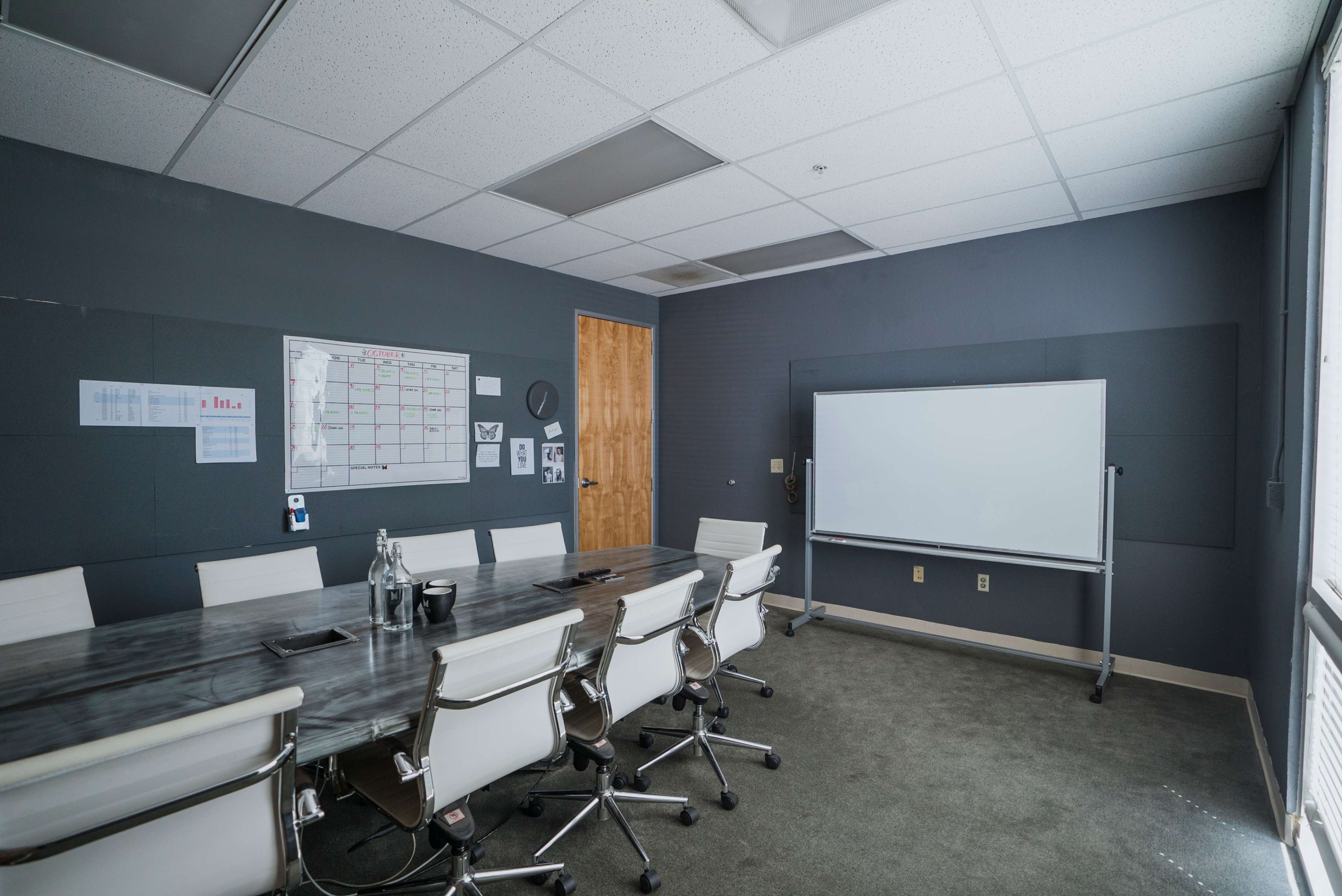 A meeting room features a long table surrounded by white chairs, a whiteboard, and a calendar on the wall.