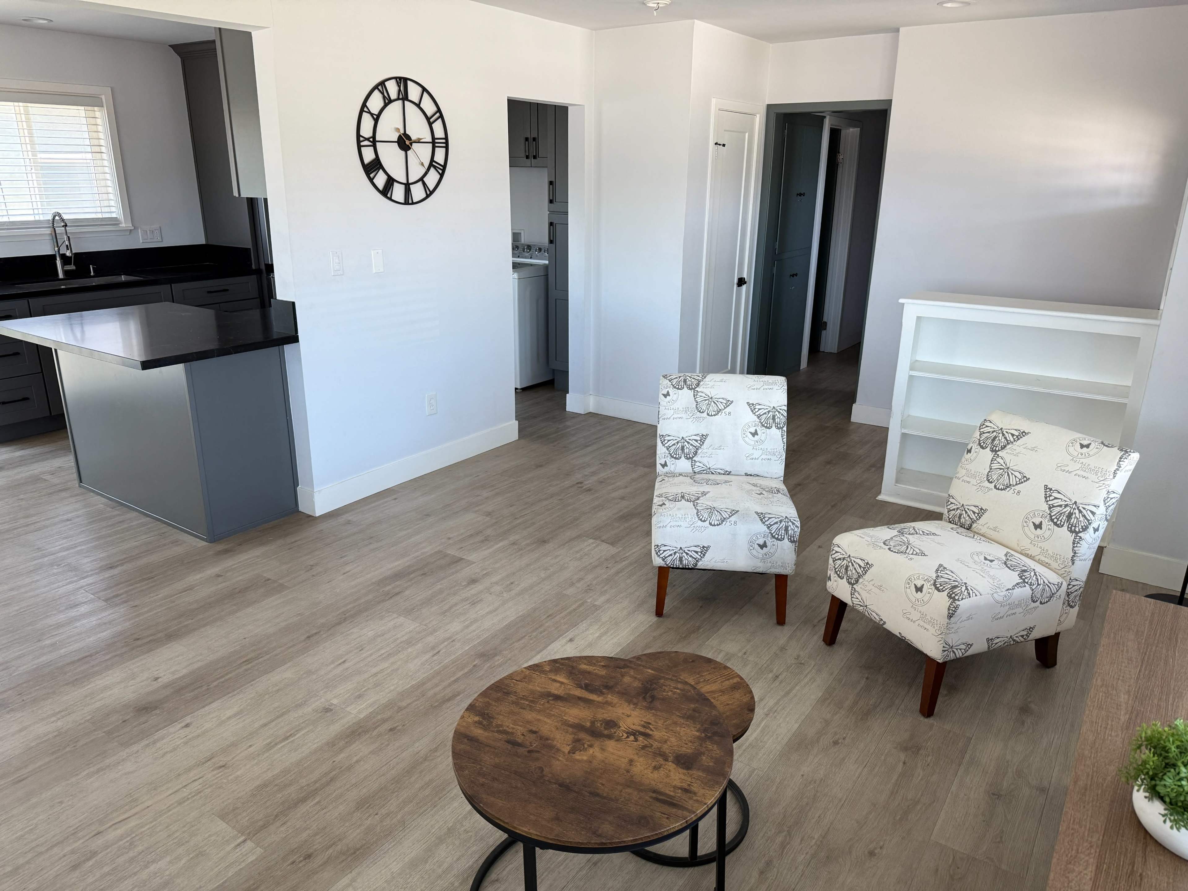 The image shows a modern living room featuring two patterned chairs, a small round table, and an open view of a kitchen area with light-colored cabinetry.