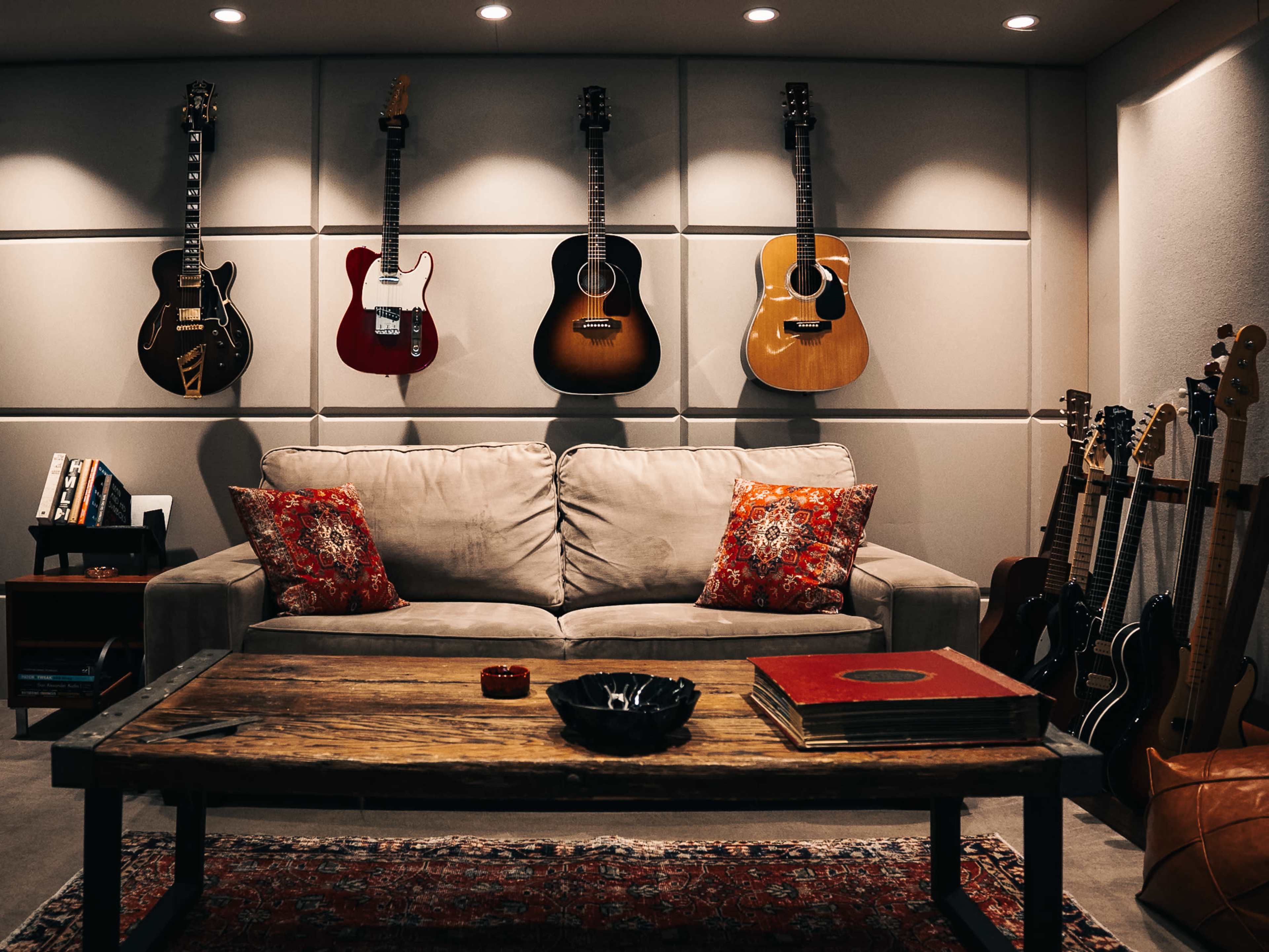 The image shows a living room featuring a beige sofa with decorative pillows, a wooden coffee table, and several guitars mounted on the wall and placed around the room.