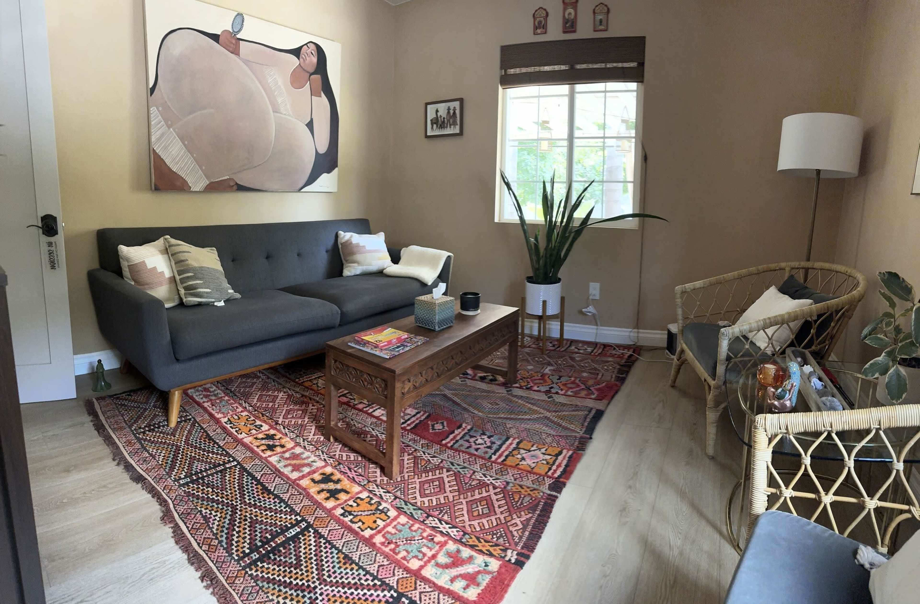 The image shows a cozy living room with a dark gray sofa, a wooden coffee table, a colorful patterned rug, and a large window that provides natural light.
