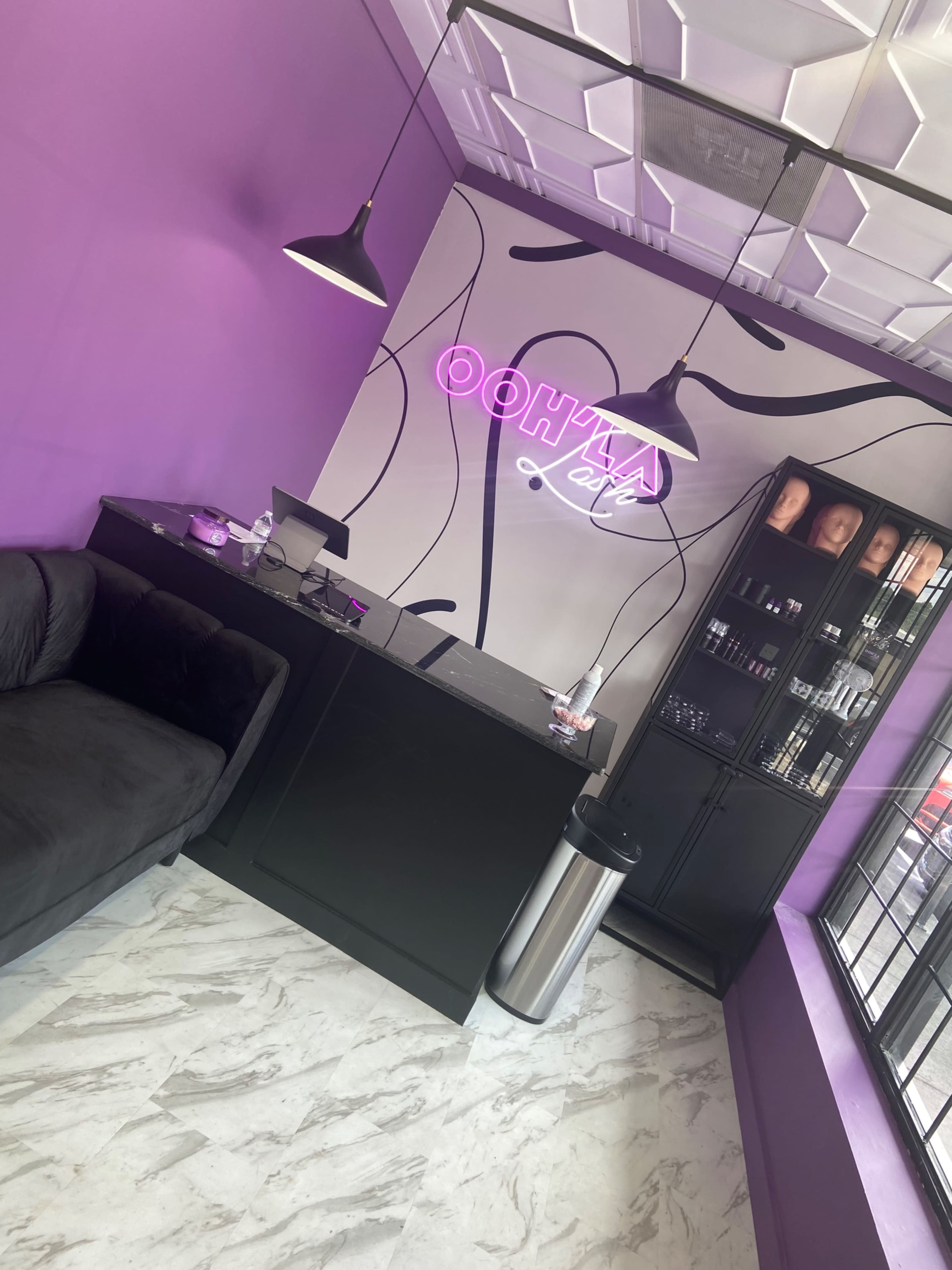The image shows a modern reception area with purple walls, a sleek black counter, a couch, and a display case showcasing hair products.