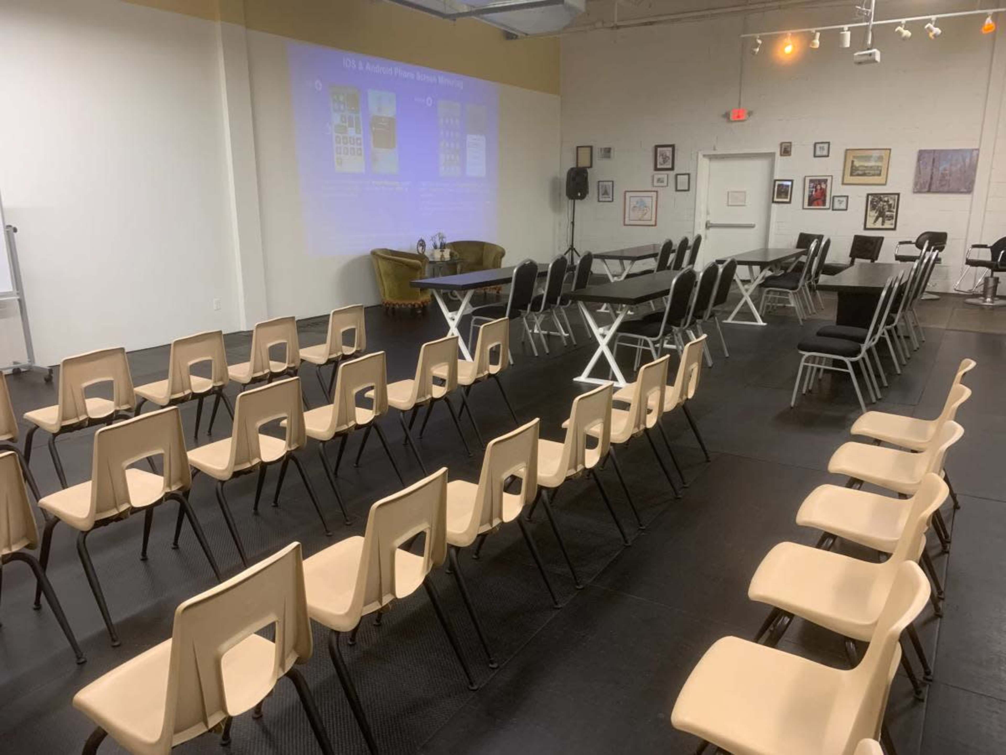 The image shows a semi-prepared event space with rows of chairs facing a presentation screen, surrounded by tables arranged for a meeting.