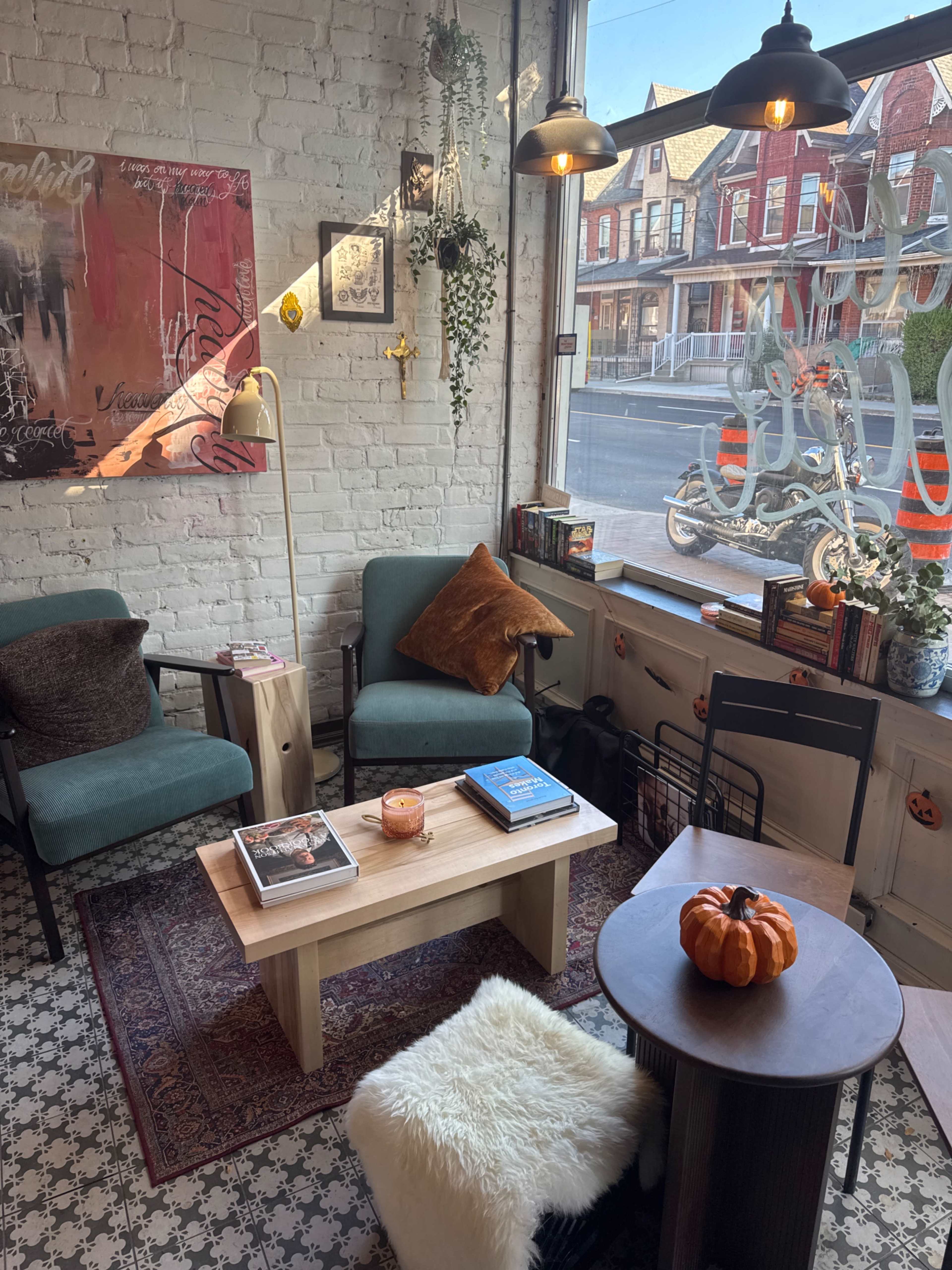 The image shows a cozy corner of a café with two green chairs, a wooden coffee table, and decorative elements like a pumpkin and books, all set against a backdrop of a street view.