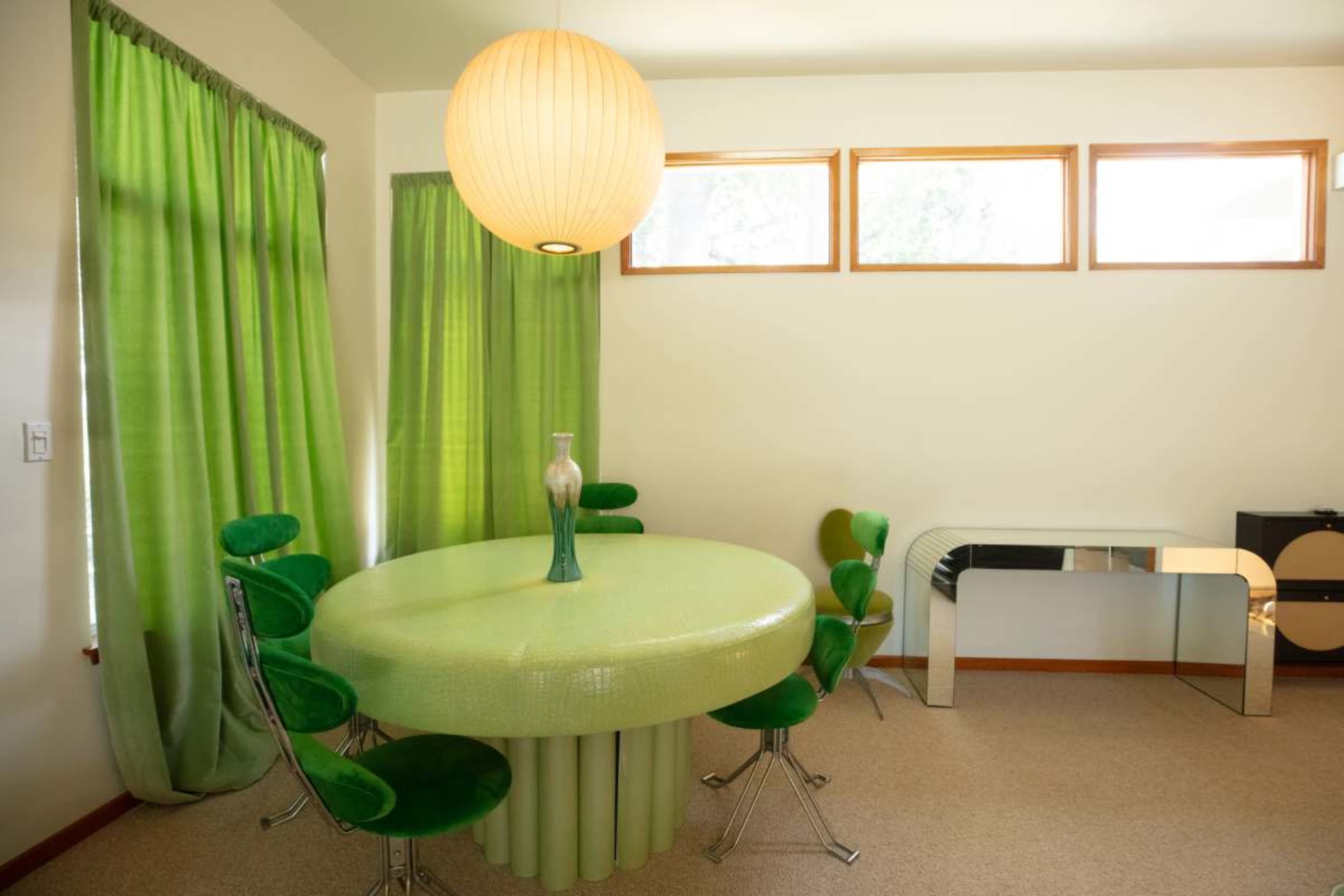 A round green table with matching chairs is set in a room with green curtains and large windows, featuring a modern console table in the corner.