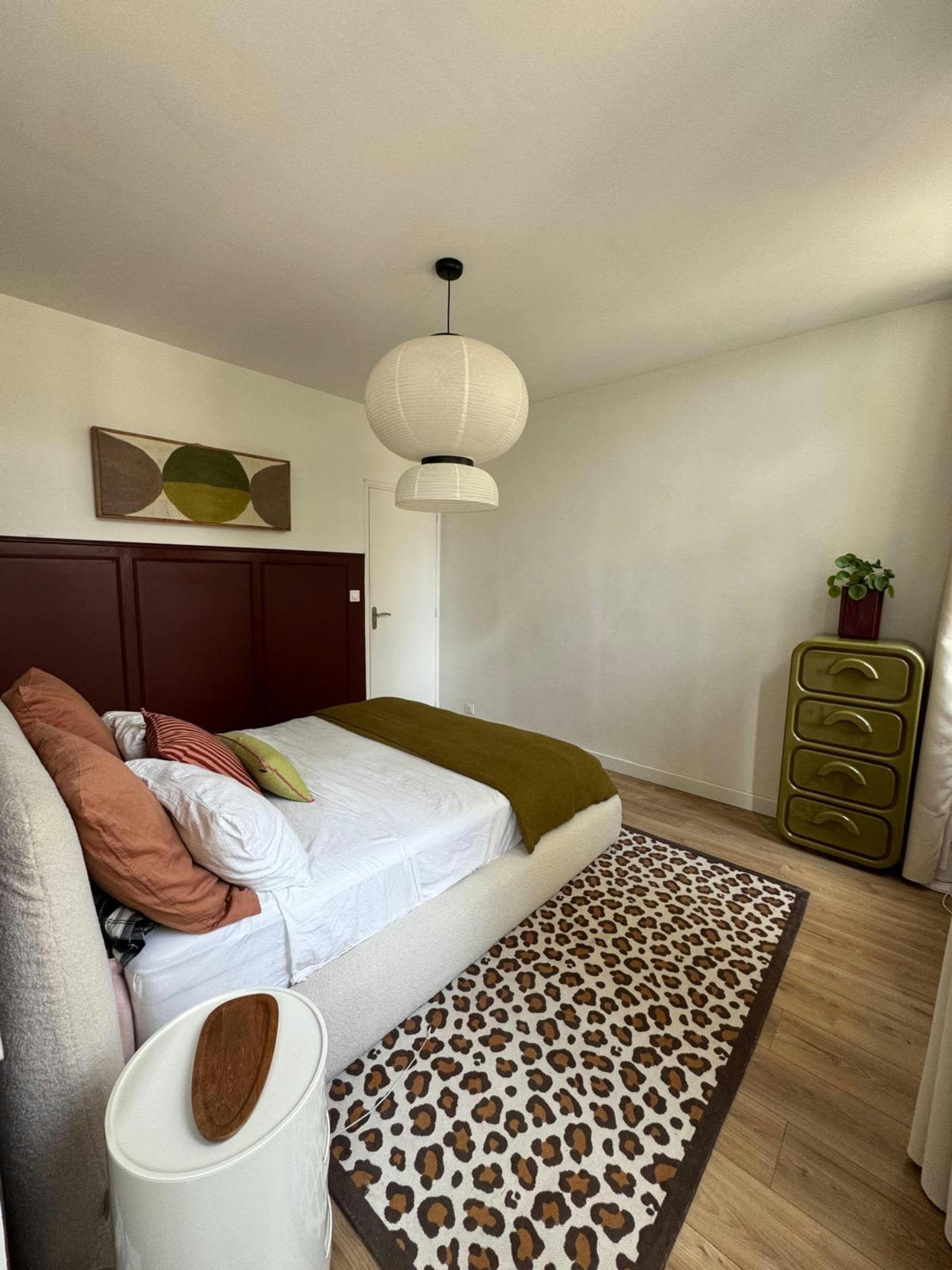 The bedroom features a bed with green and white linens, a leopard-print rug, and a decorative chest of drawers against a light-colored wall.