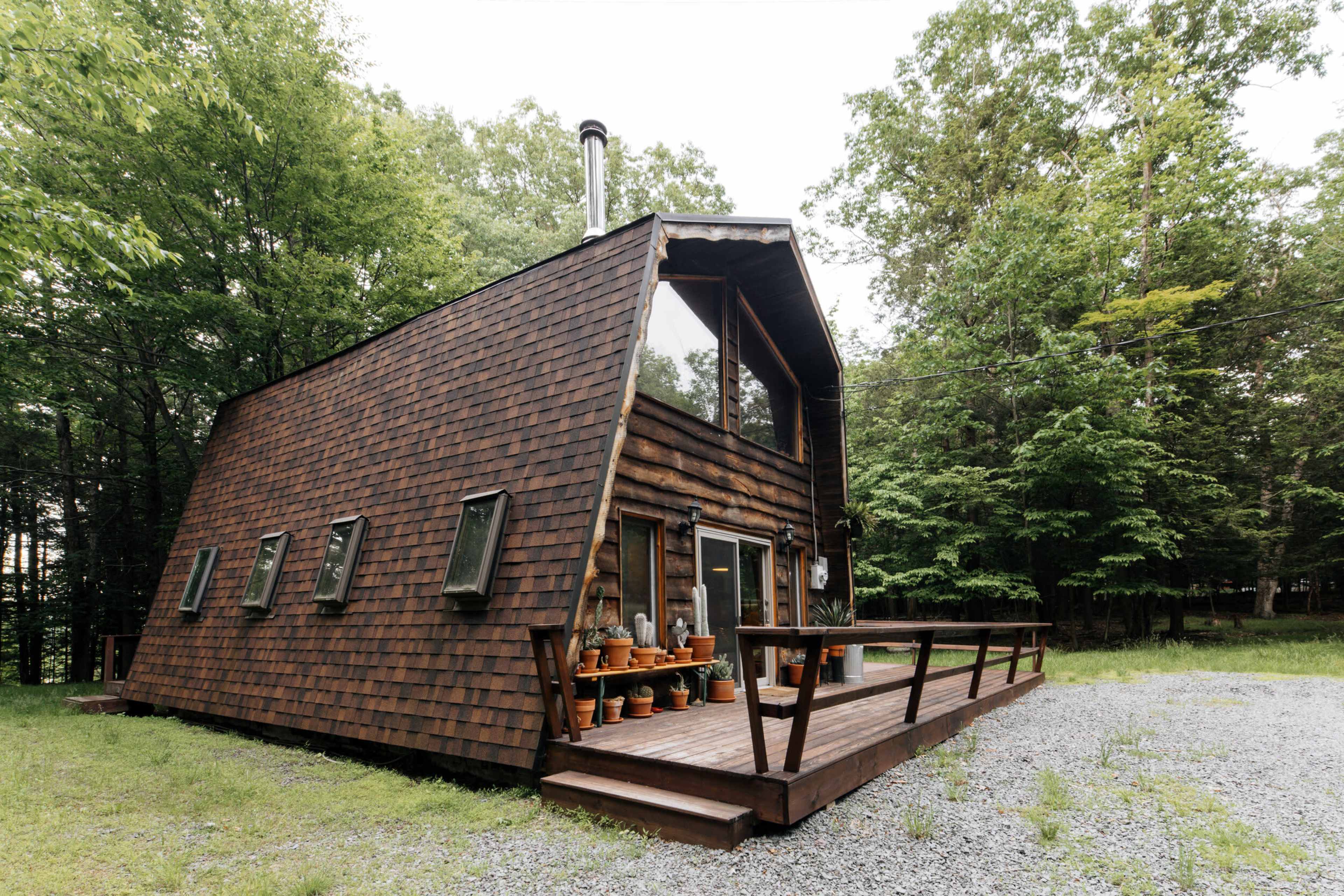 A wooden, A-frame house with a sloped roof is surrounded by trees and features a porch with potted plants.