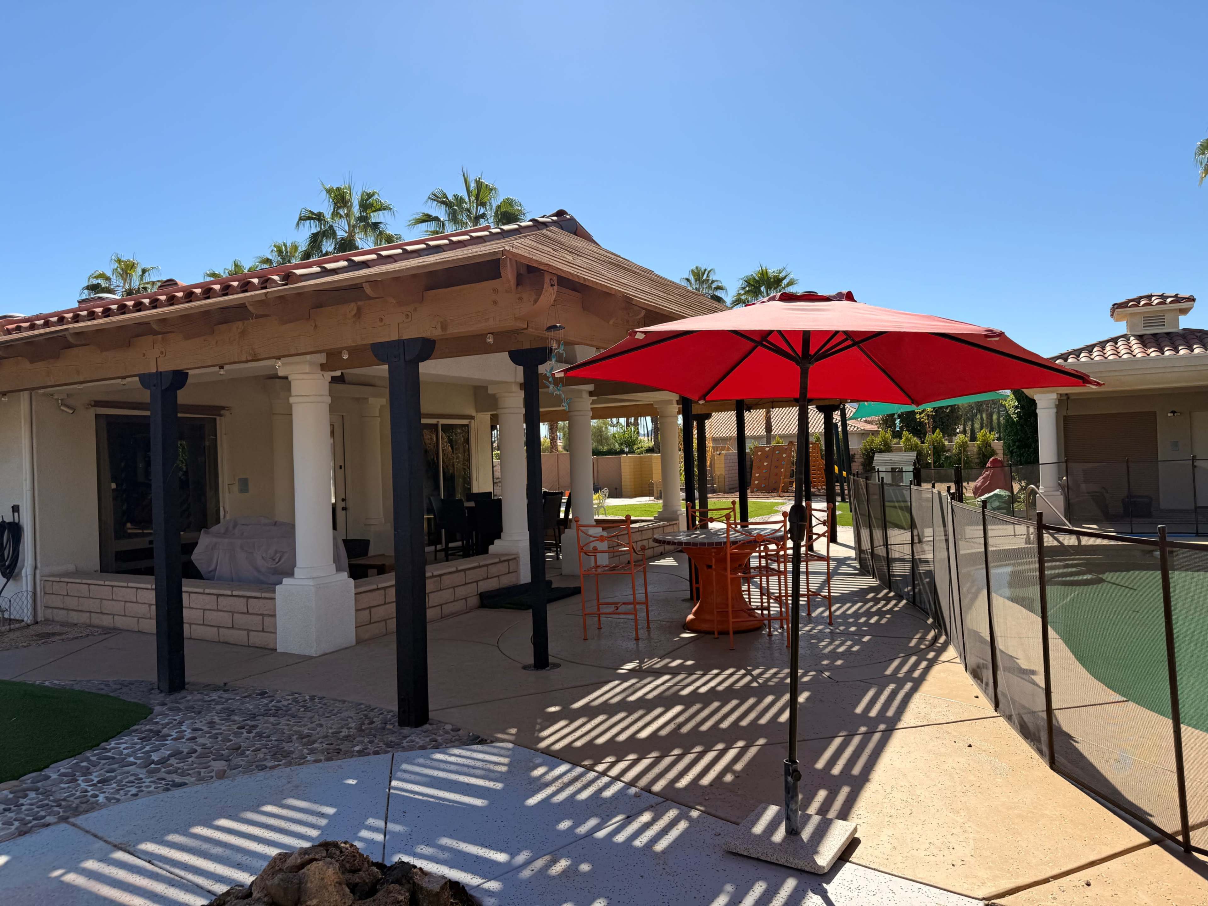 A red umbrella shades a dining area next to a poolside patio with a covered seating area and a fenced landscape.