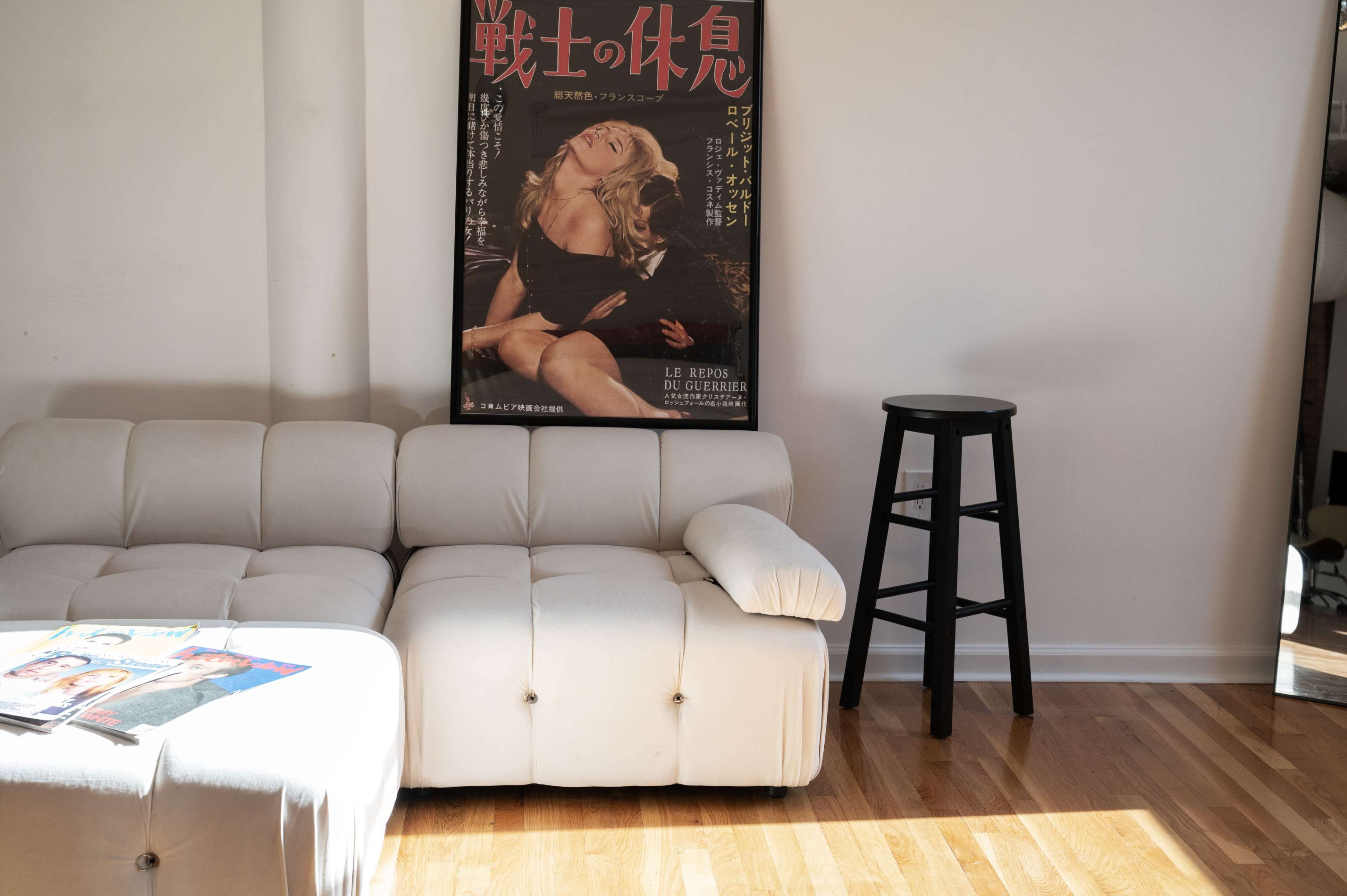 The scene features a light-colored couch positioned against a wall adorned with a large vintage movie poster, alongside a black stool and a magazine on a coffee table.