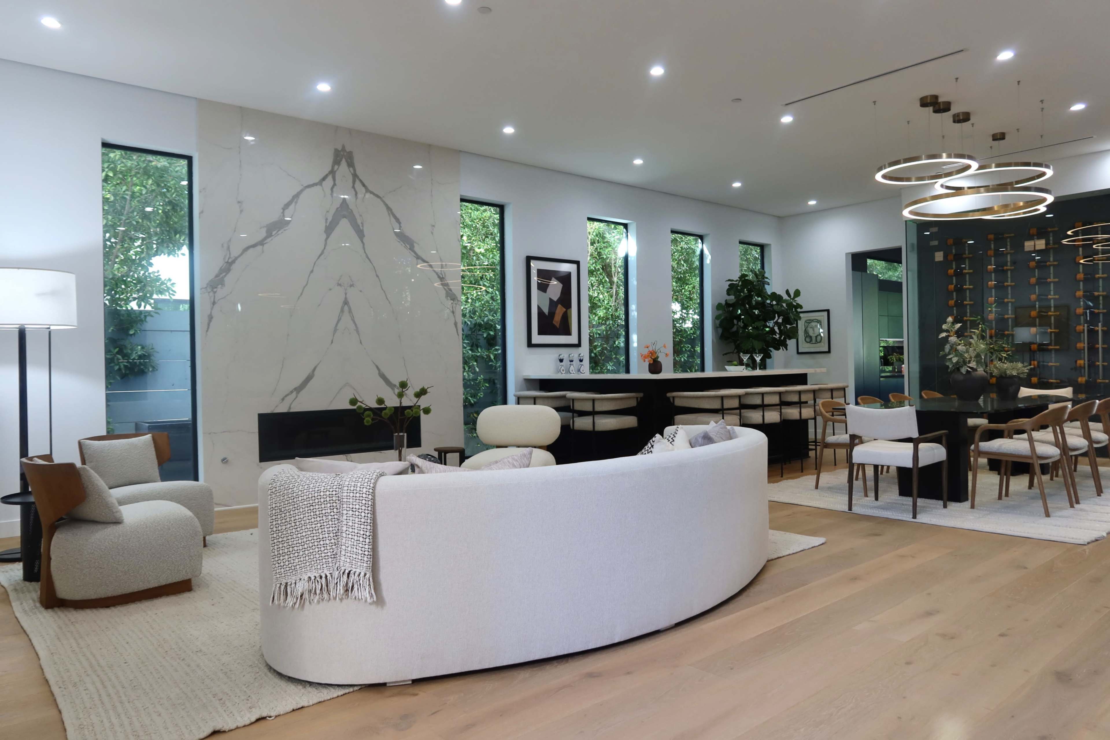 The image shows a modern living space with a curved sofa, a sleek fireplace, large windows, and an open dining area featuring a wooden table and stylish chairs.
