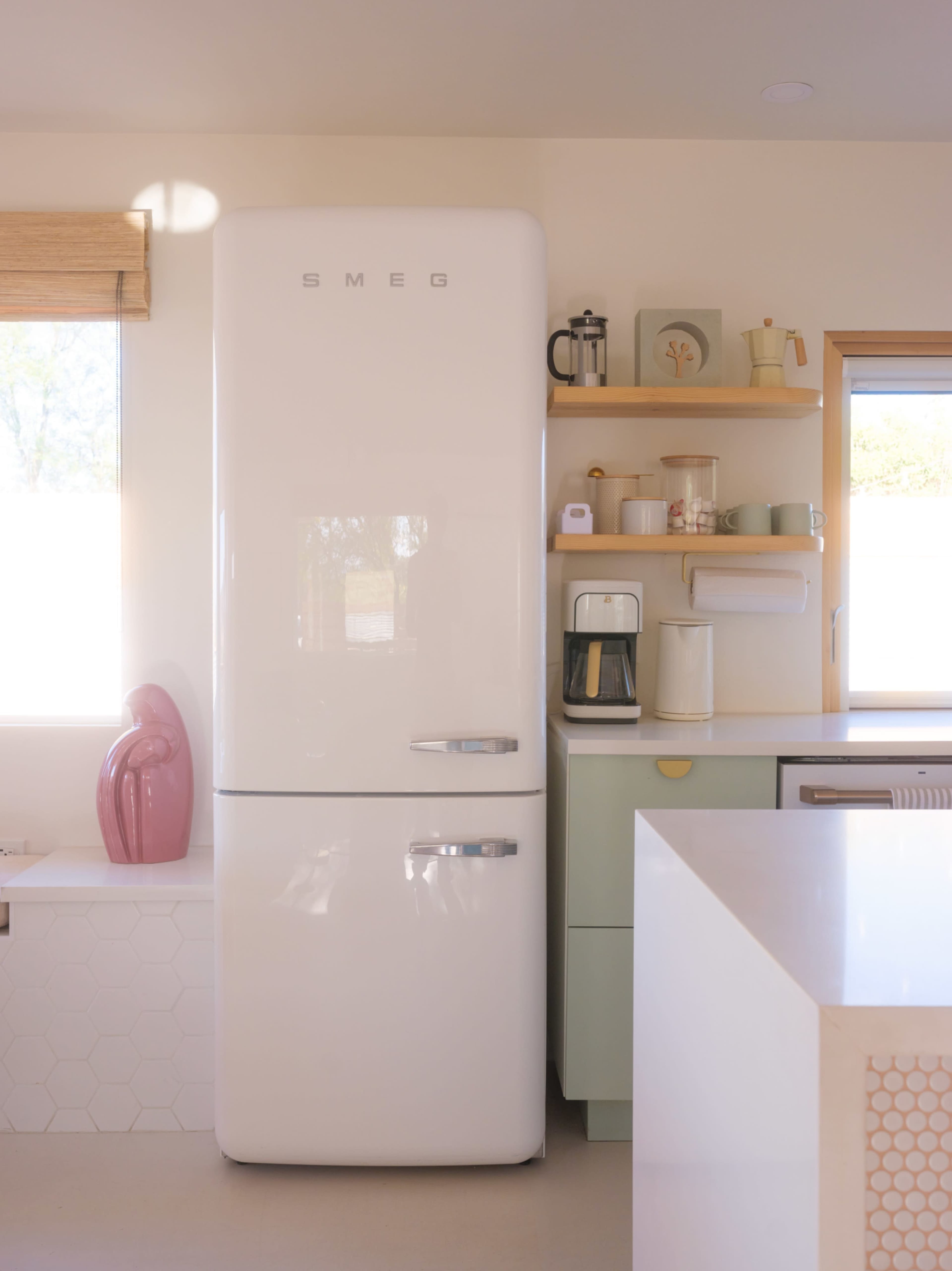 A white SMEG refrigerator stands next to a shelf displaying a coffee maker and various kitchen items in a bright, modern kitchen.