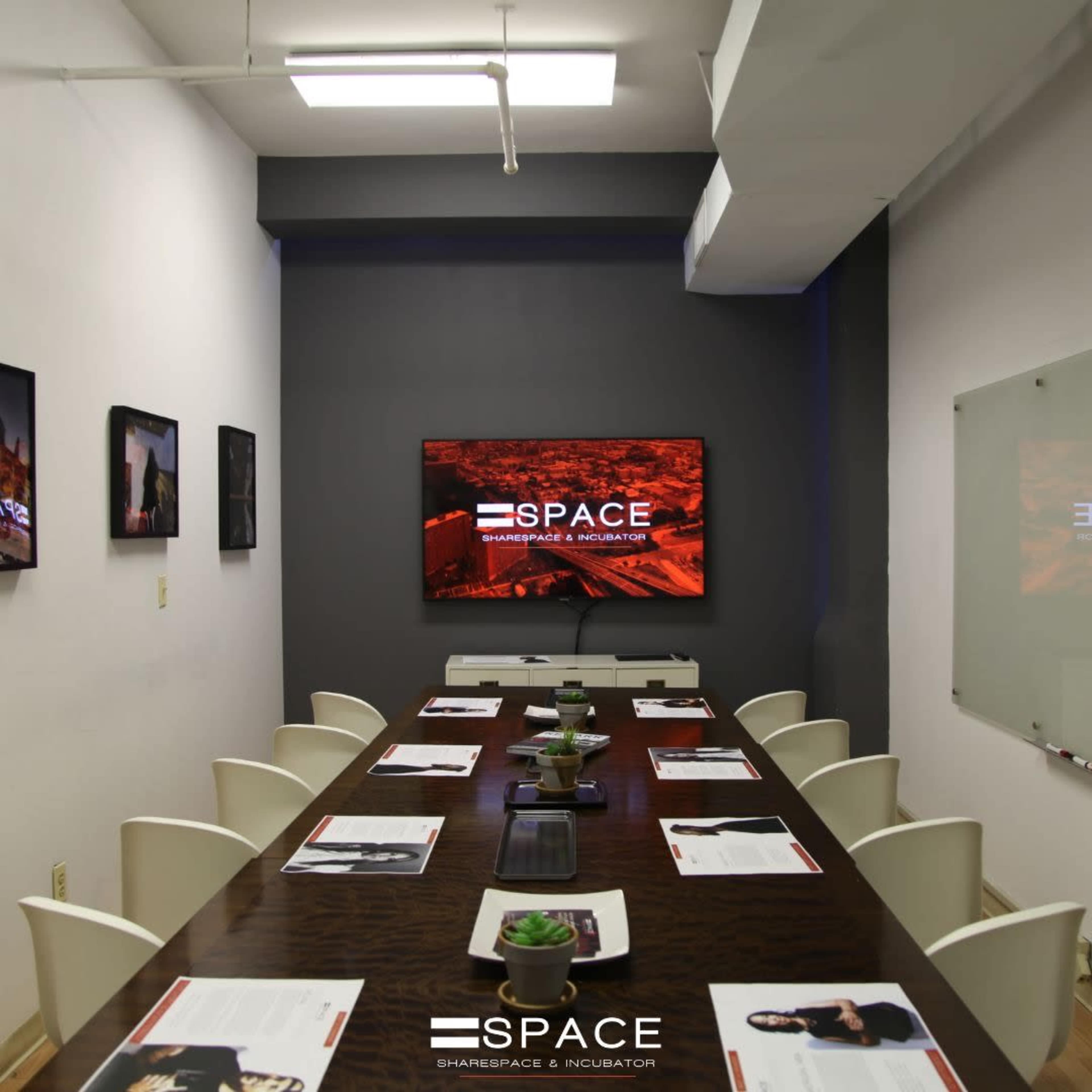 A modern conference room features a long wooden table with chairs, a large television displaying a logo, and placemats set for a meeting.