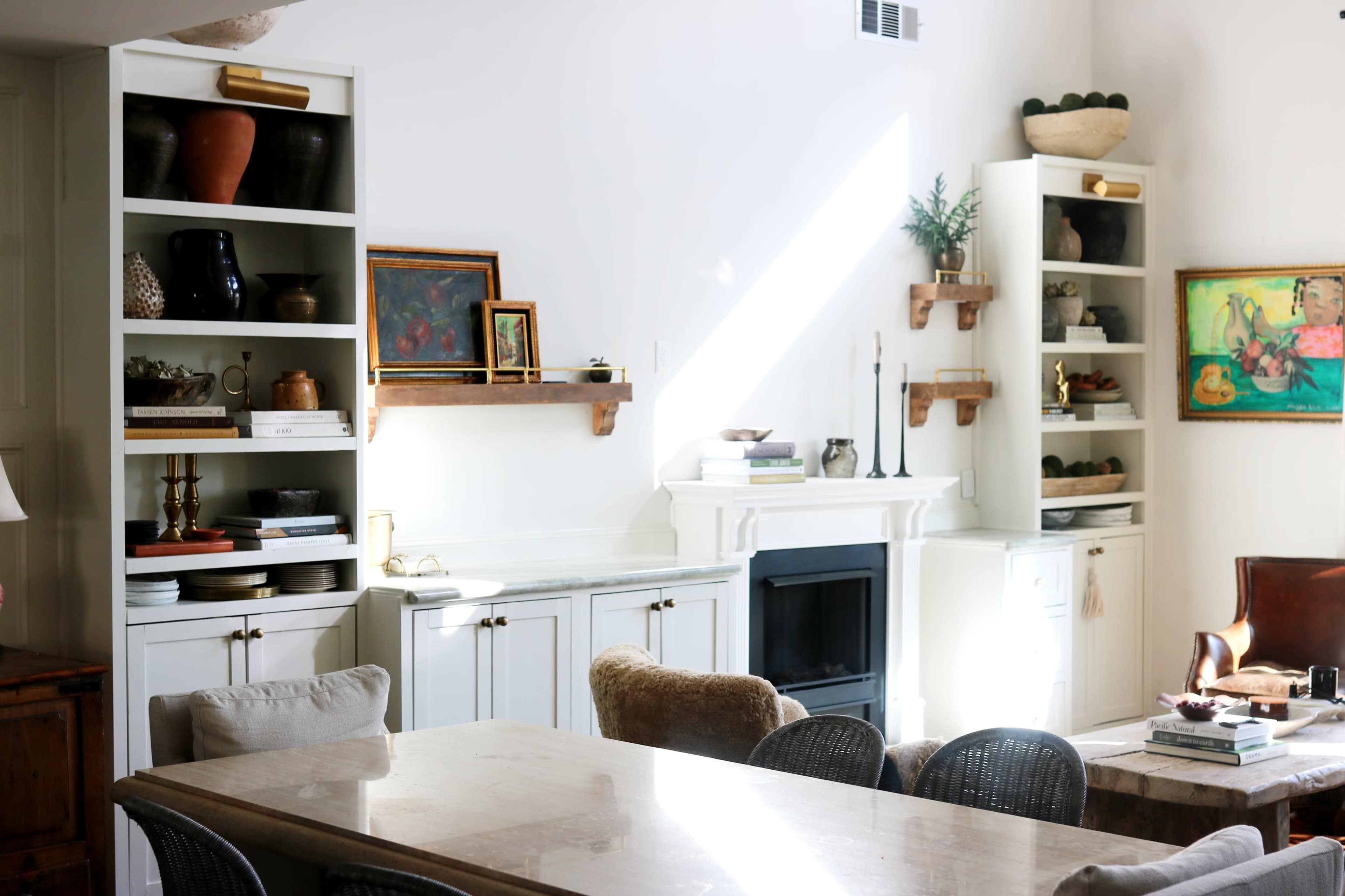 The image shows a bright living space featuring white shelves filled with decorative items, a central dining table, and a fireplace with a mantel.