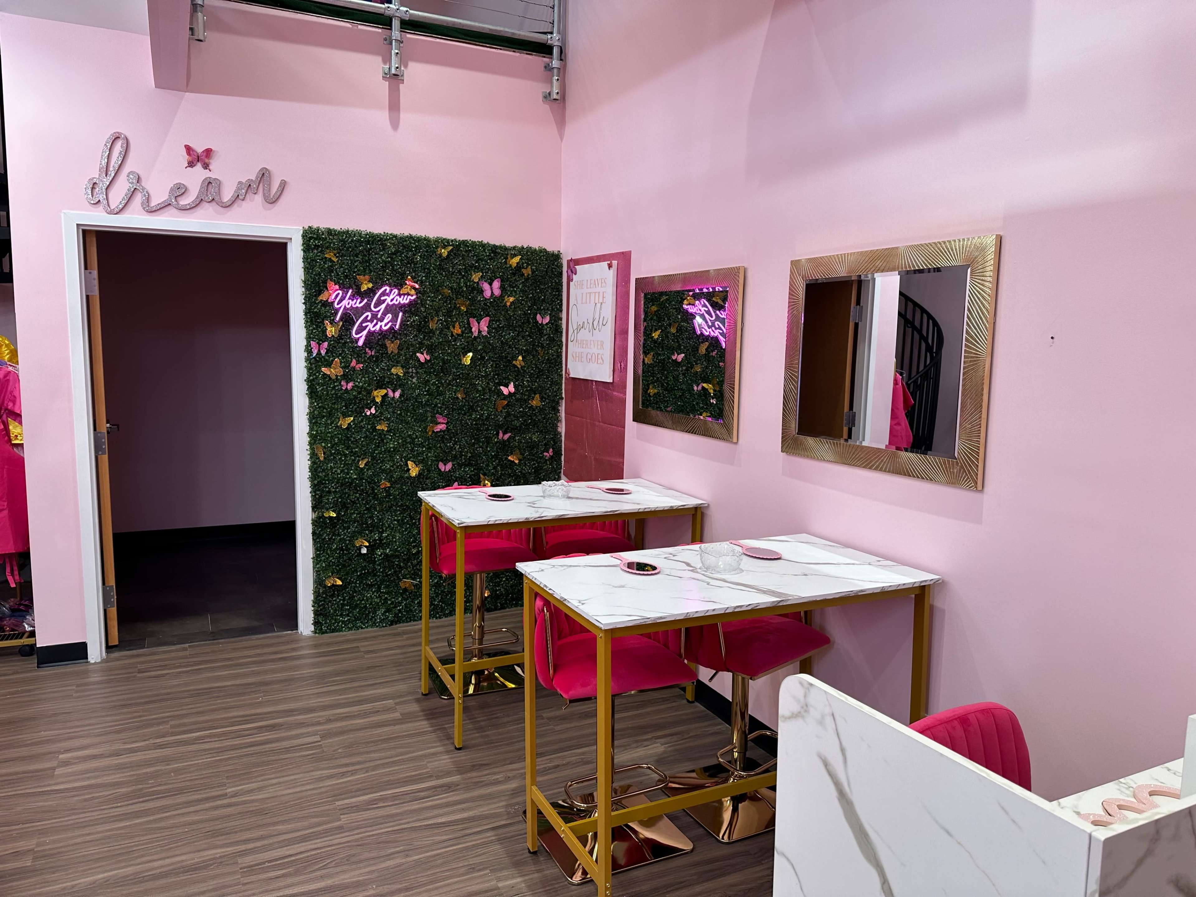 The image shows a pink-themed interior featuring two marble-topped tables with gold legs and pink chairs, complemented by a green wall adorned with artificial plants and butterflies.