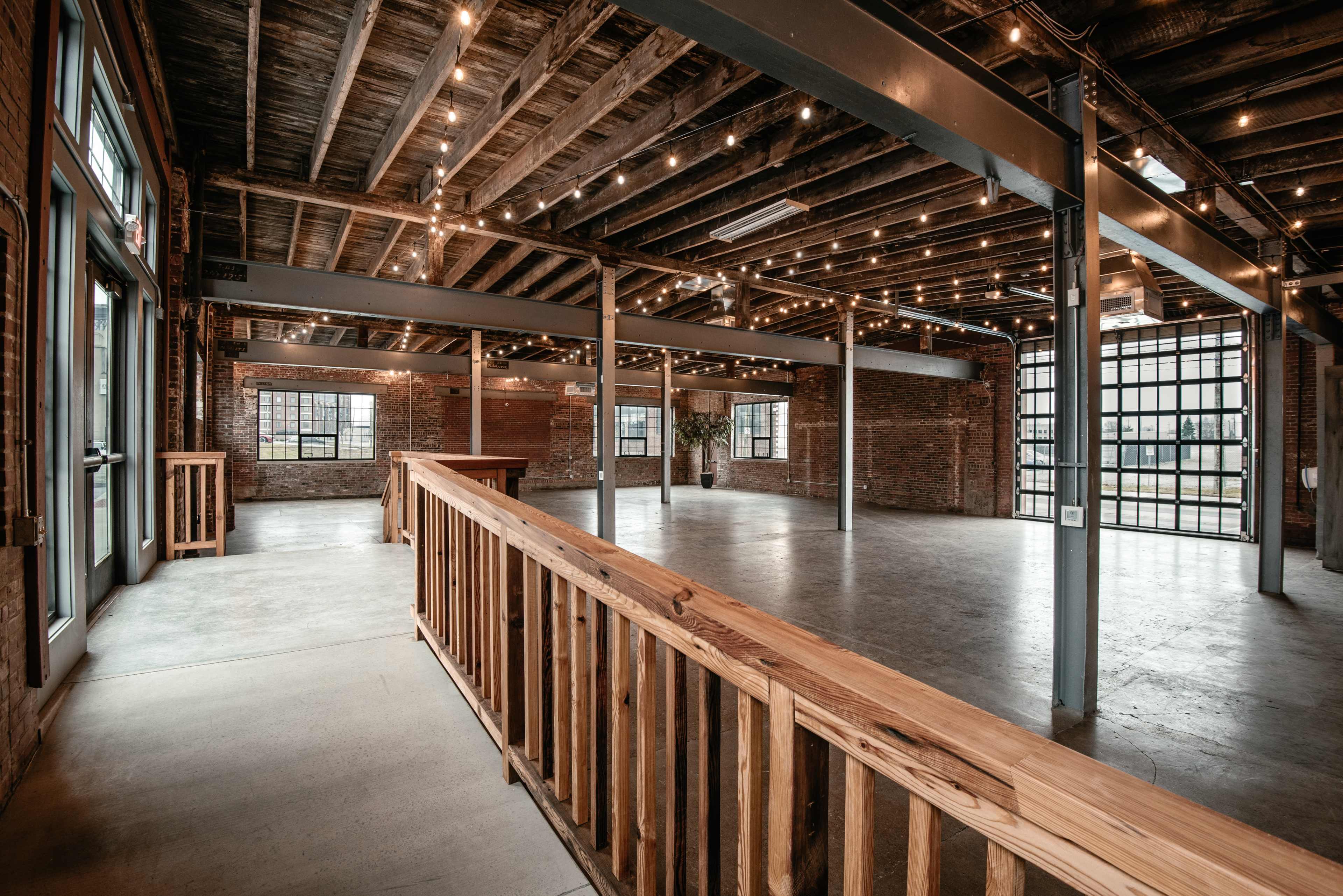 A spacious, industrial-style venue with exposed brick walls, wooden beams, and a polished concrete floor, illuminated by string lights.
