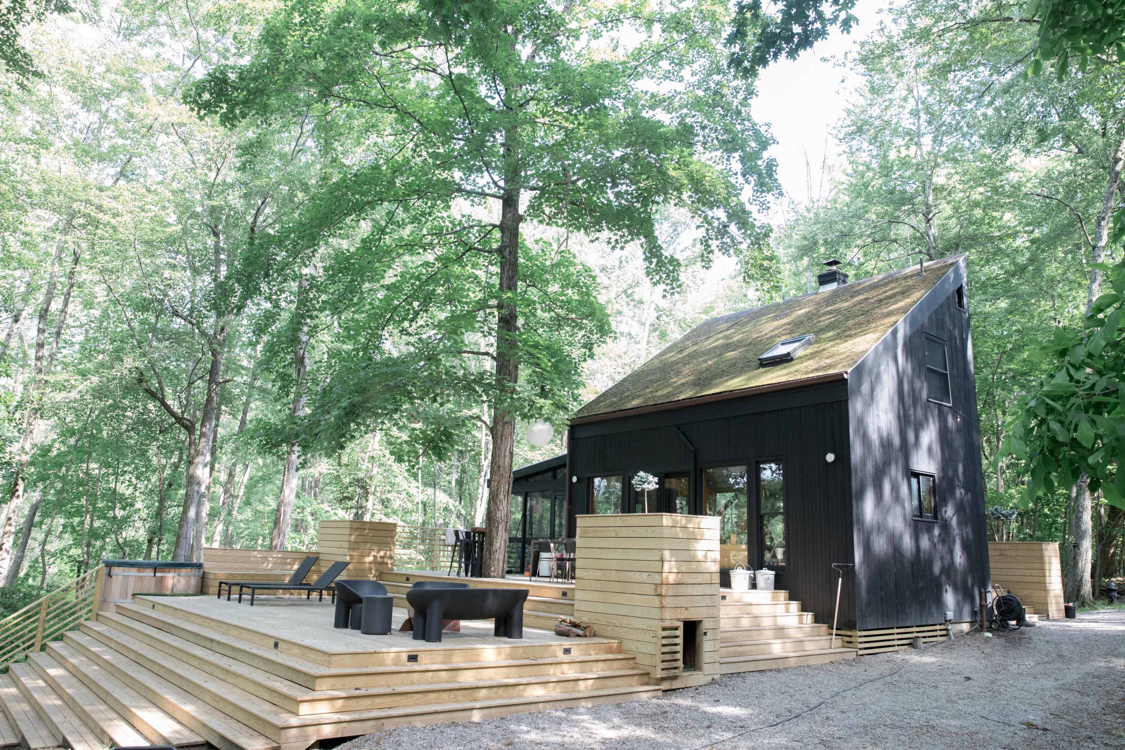 A modern black house with a sloped roof is surrounded by green trees and features a wooden deck with seating areas.