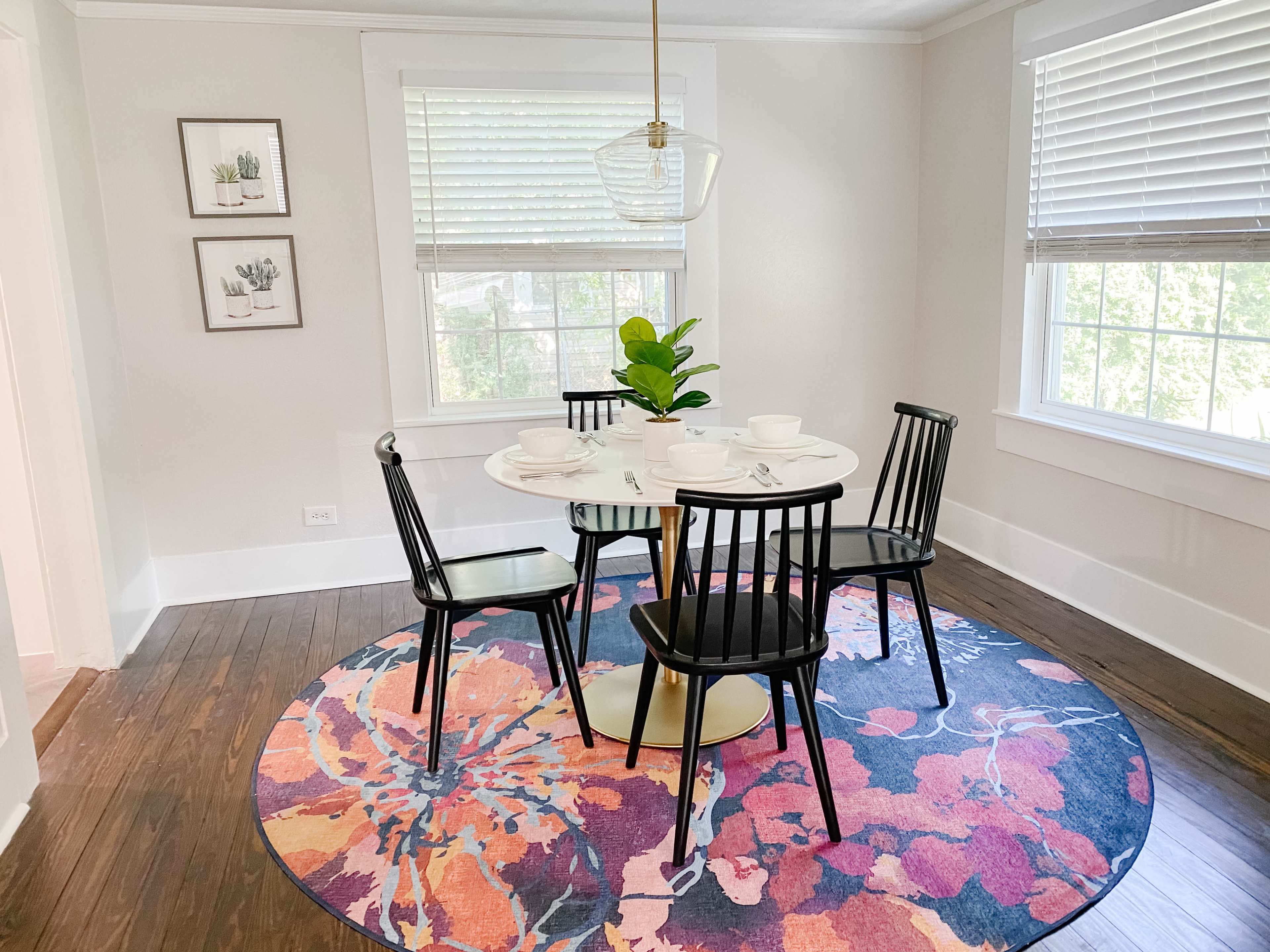 A round dining table set with white dishes and four black chairs is placed on a floral area rug in a well-lit dining room with two windows.