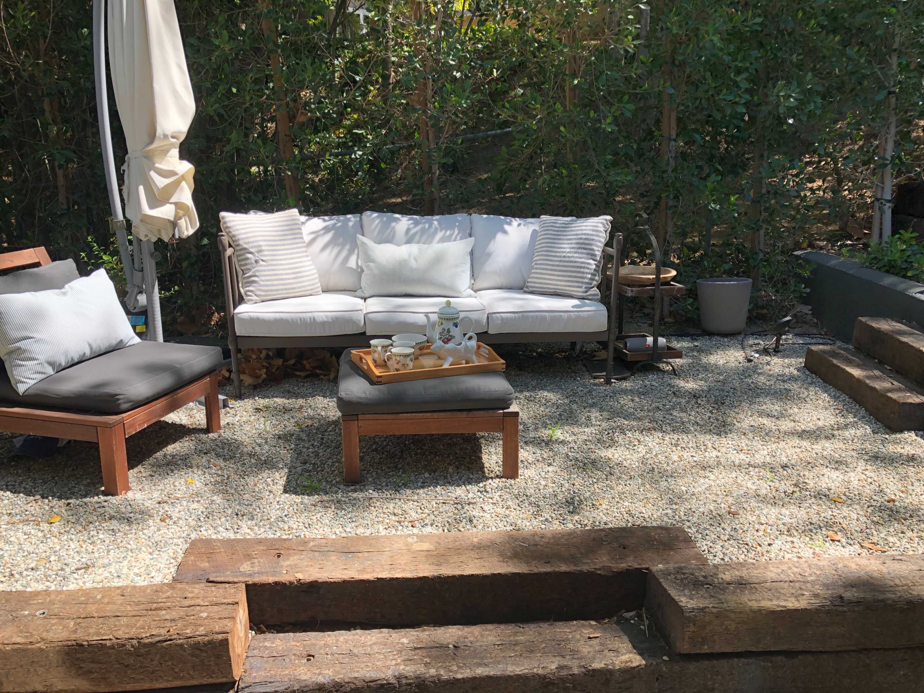 A seating area with a white sofa and a coffee table is set on gravel, surrounded by greenery and wooden steps.