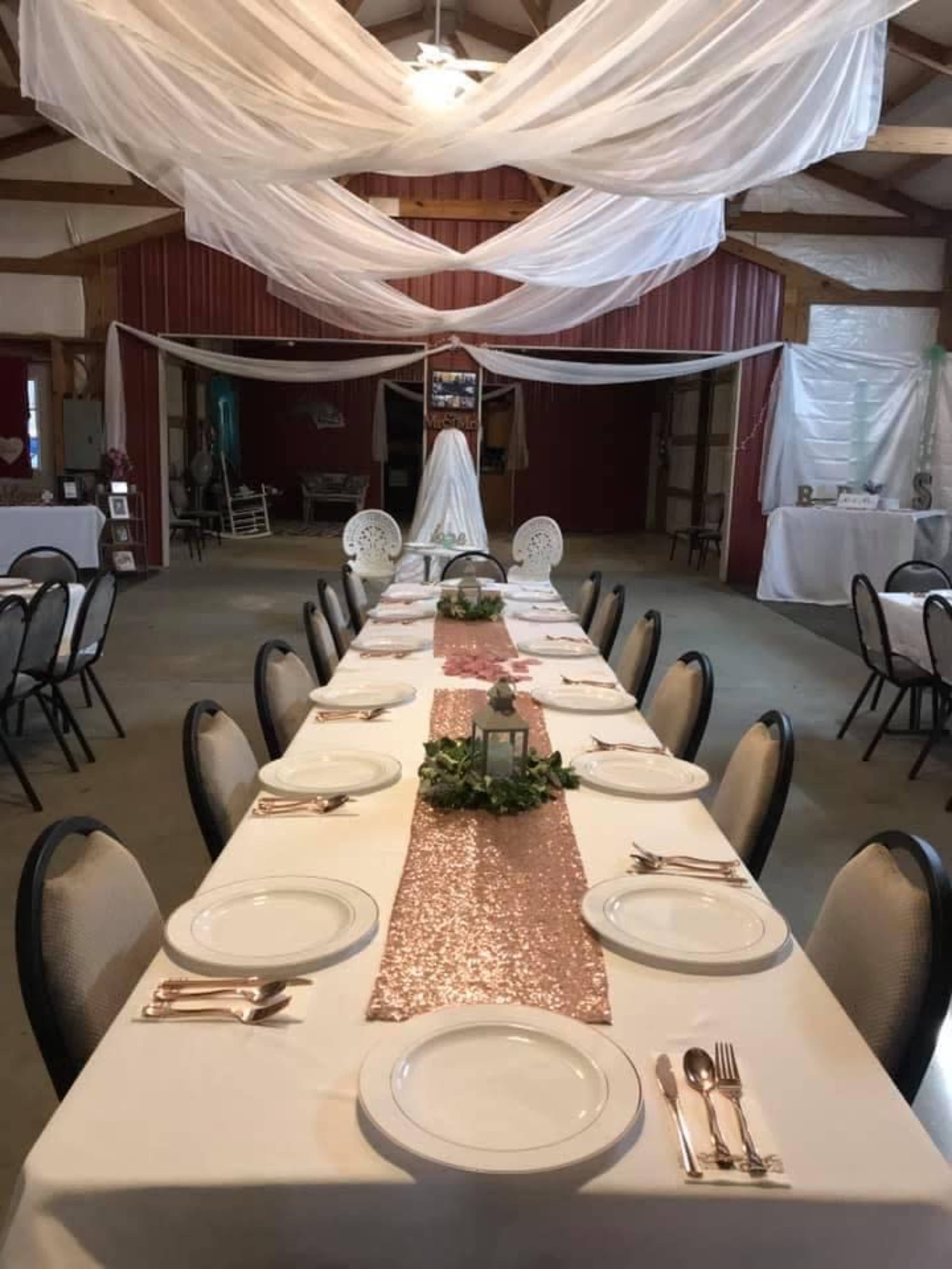 A long banquet table is set with white plates and silverware, adorned with a sparkling rose gold runner and greenery, under draped fabric in a rustic venue.