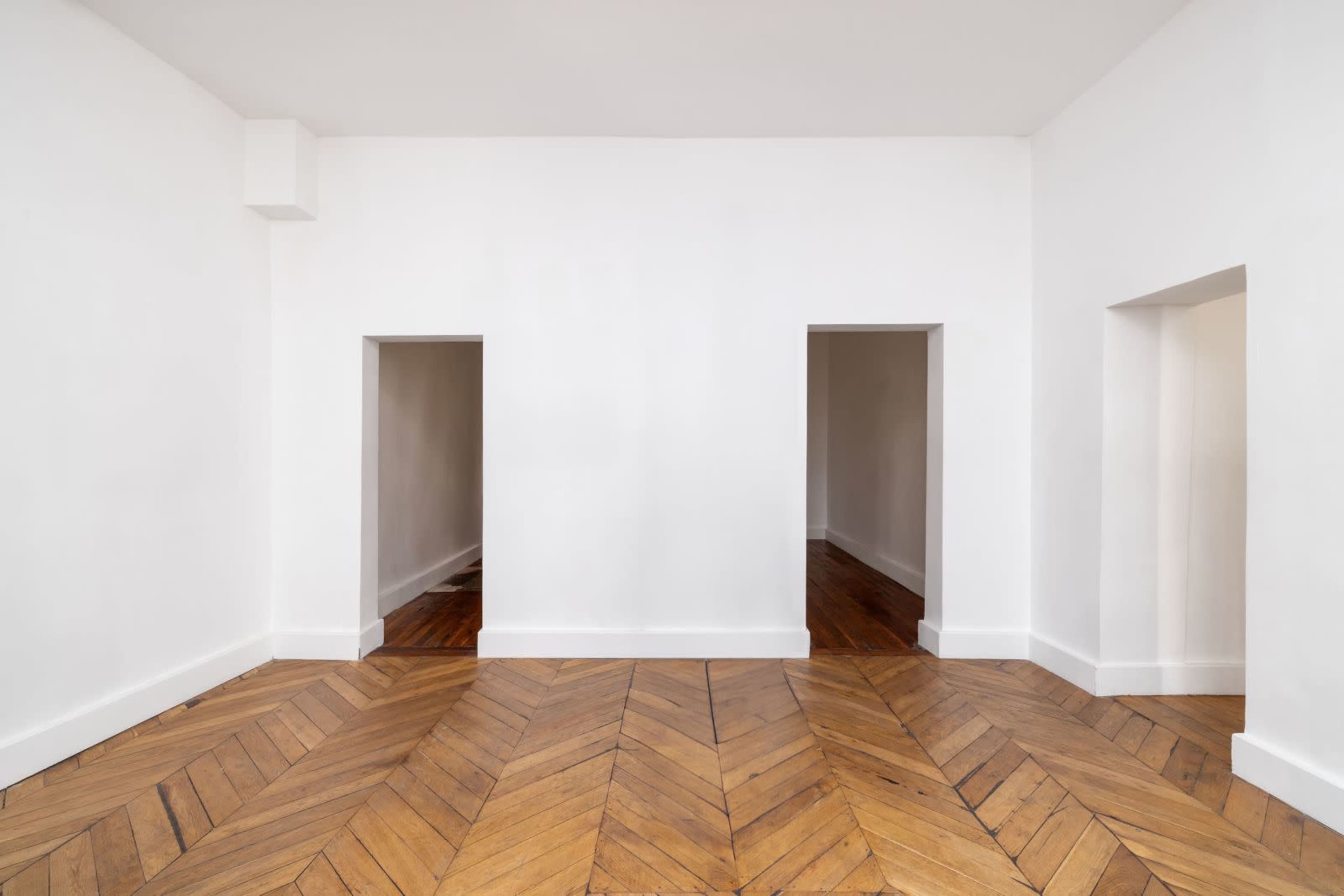 The image depicts a minimalistic room with white walls, a wooden herringbone floor, and two empty doorways on opposite sides.
