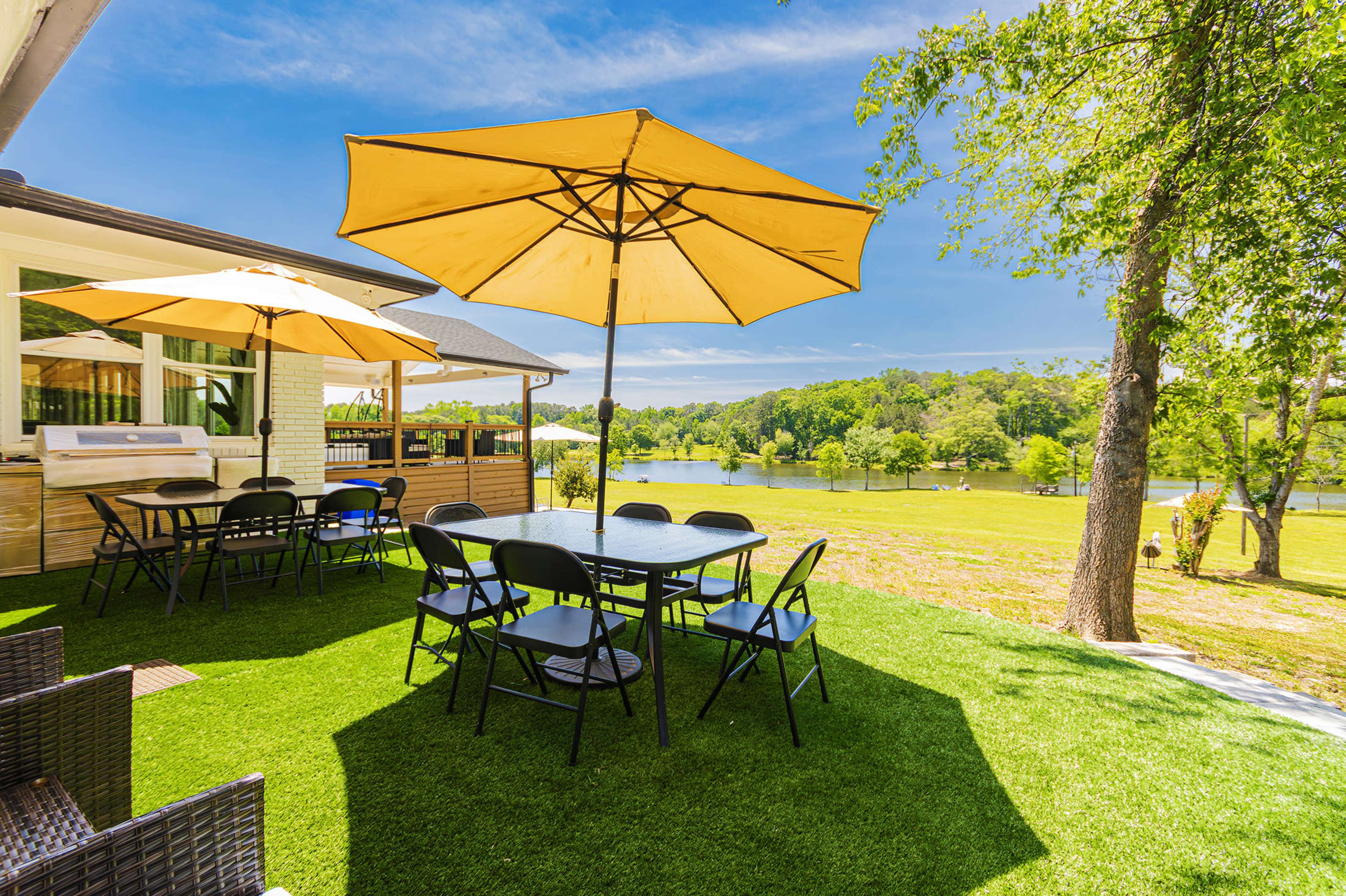 The image shows a patio area with black folding chairs and tables under yellow umbrellas, overlooking a green lawn and a calm body of water.