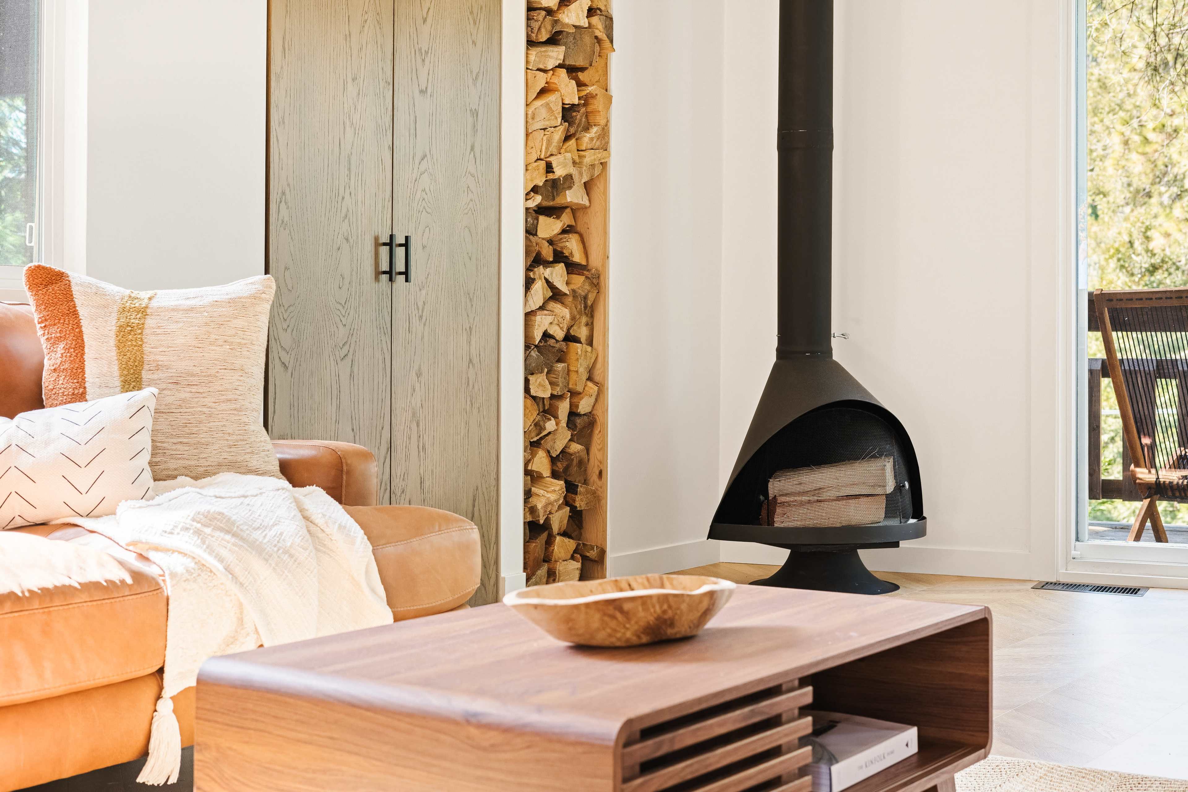 A cozy living room featuring a leather sofa, a wooden coffee table, a modern fireplace, and neatly stacked firewood beside an oak cabinet.