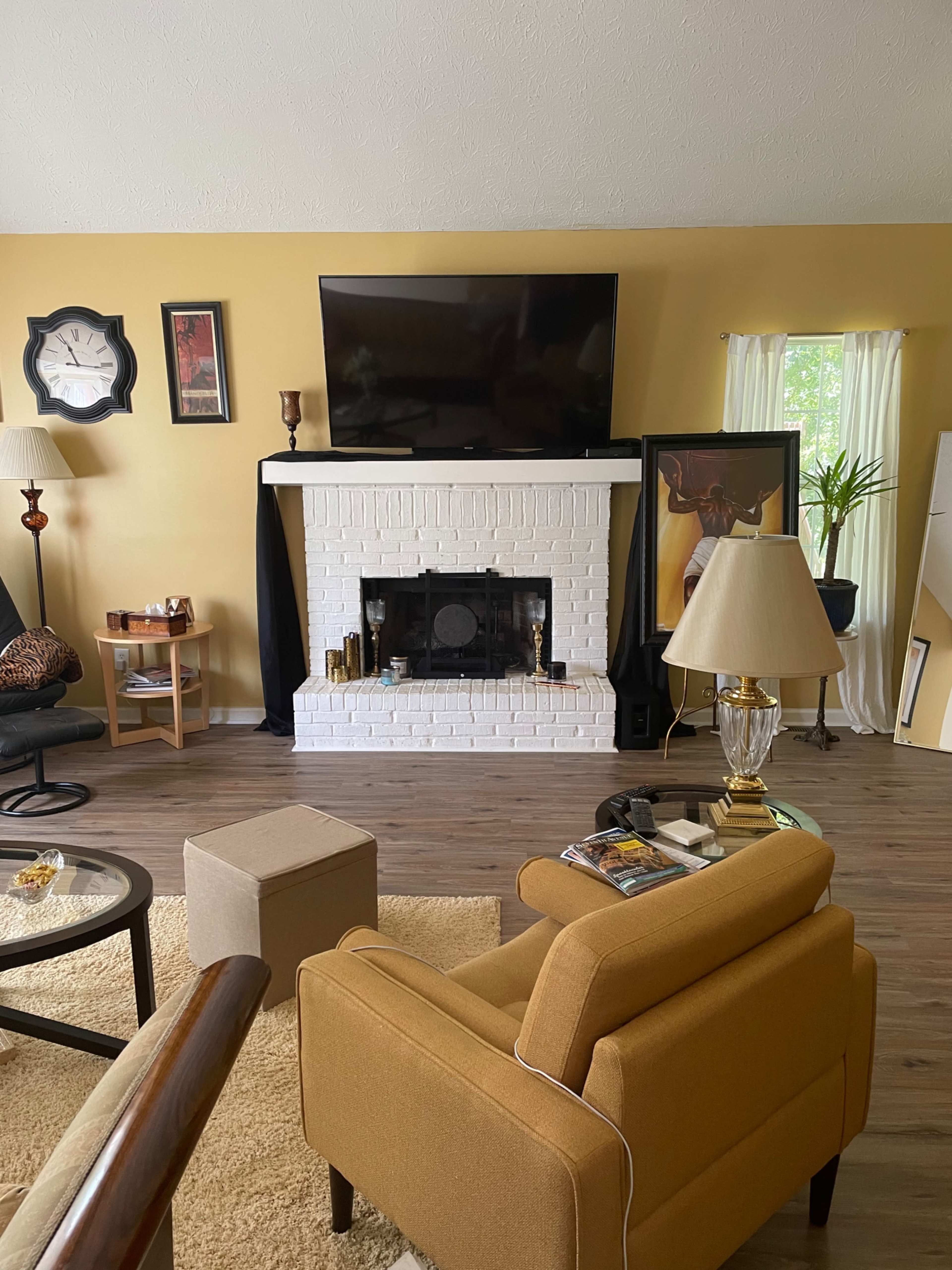 The living room features a yellow wall, a white brick fireplace with a television mounted above, and a variety of furniture including a brown armchair and a lamp.
