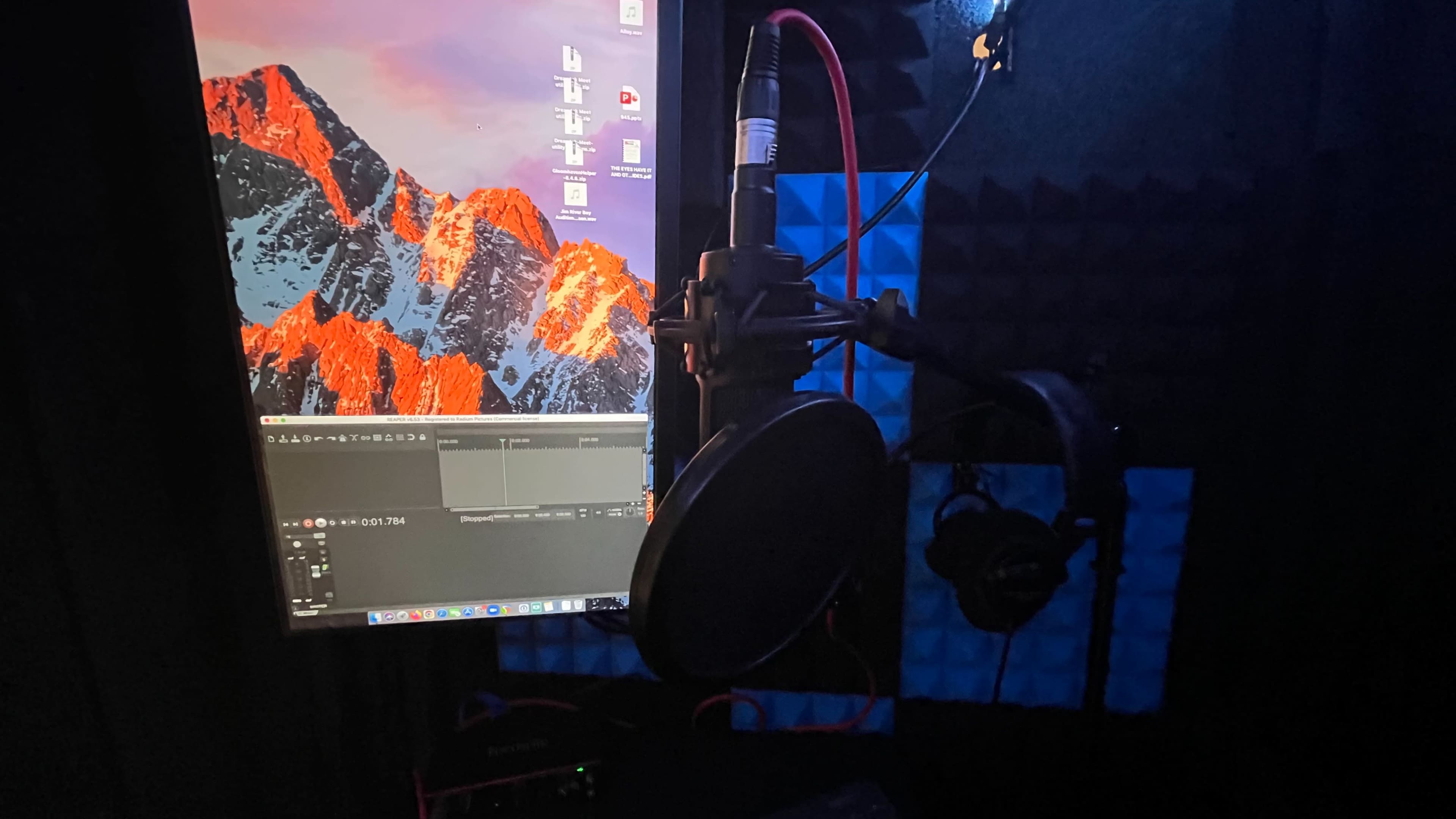 A recording setup featuring a microphone mounted on a stand next to a computer monitor displaying editing software, all situated within a soundproof room.