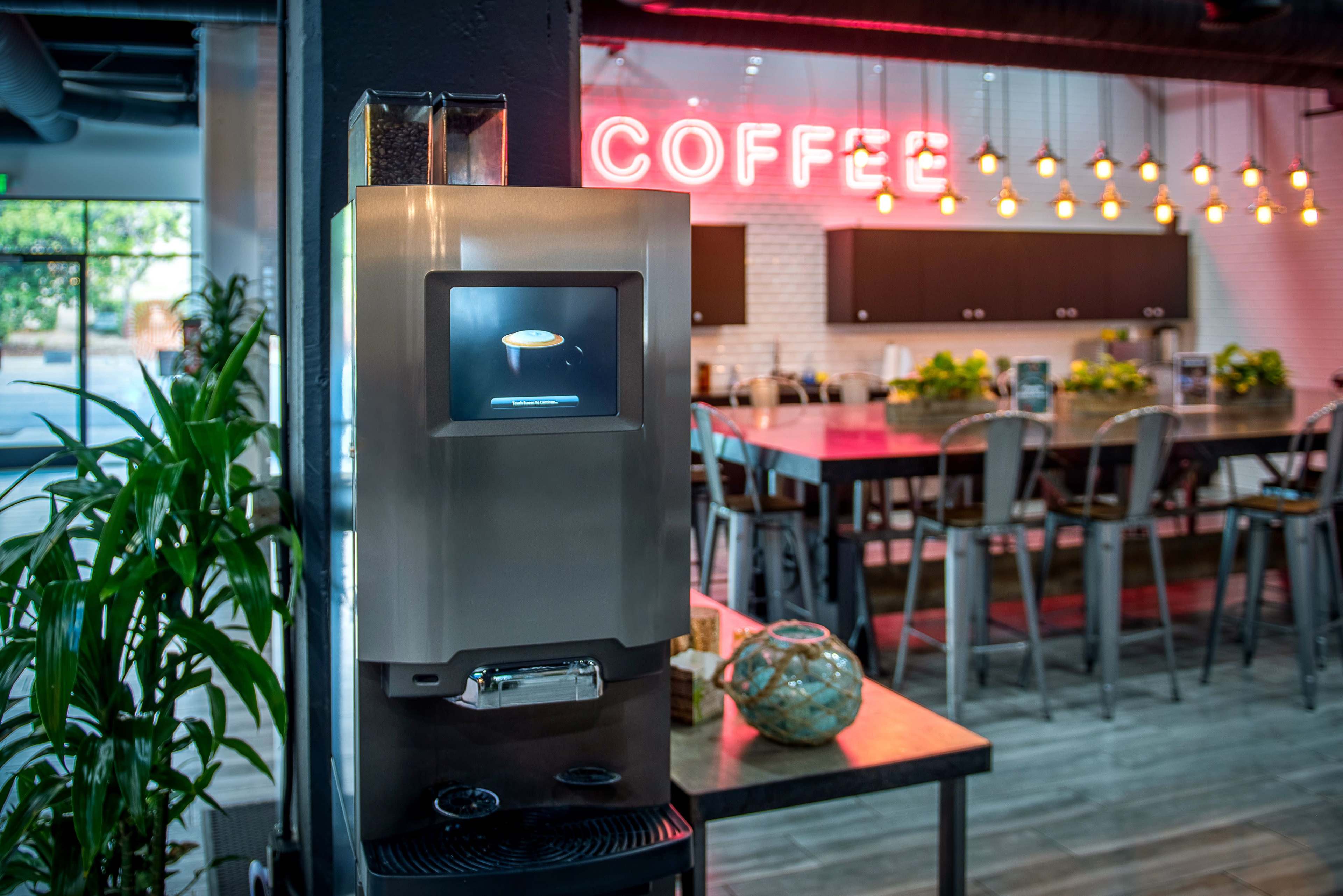 A modern coffee machine stands beside a large dining table in a cafe with illuminated "COFFEE" signage in the background.