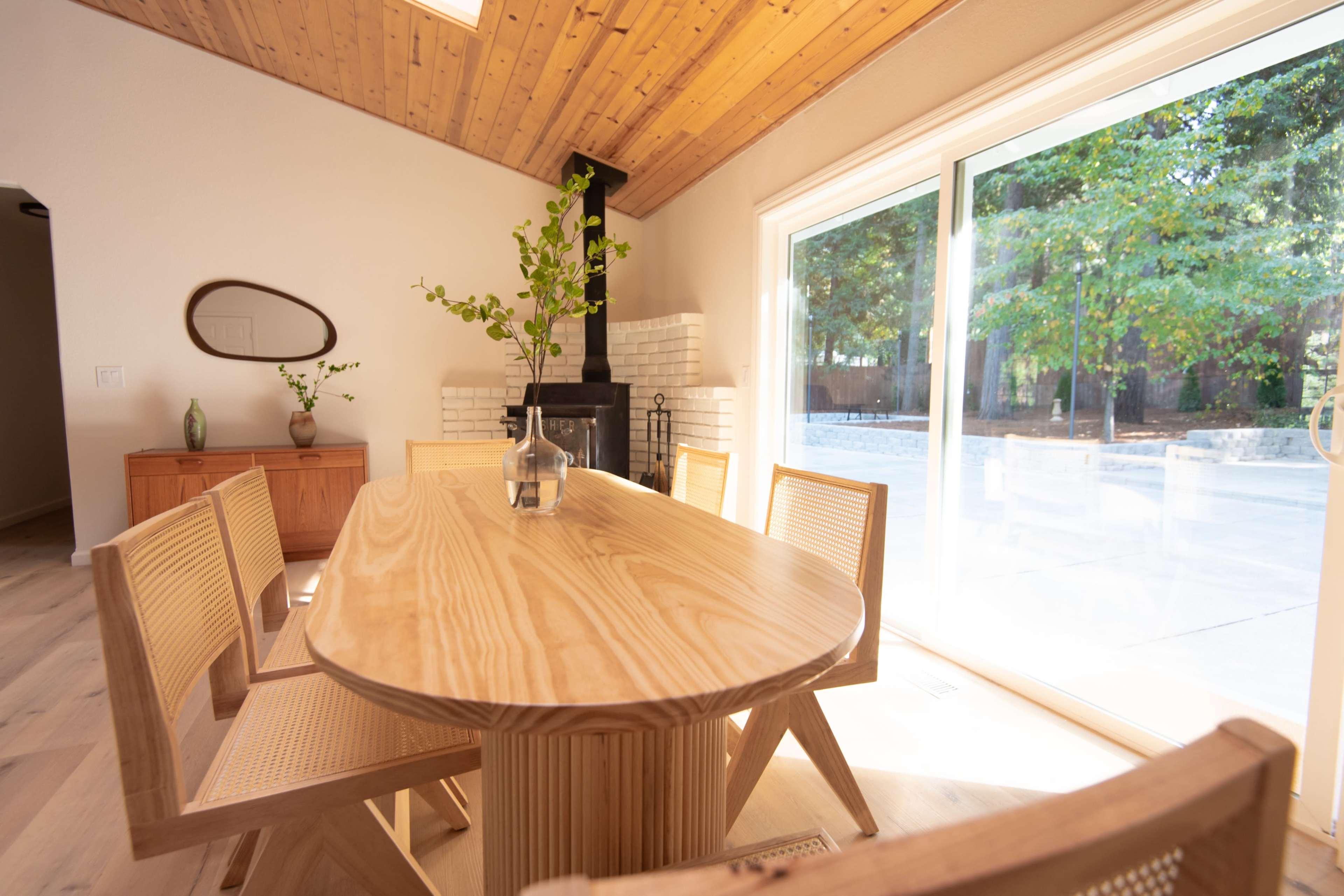 A wooden dining table with six chairs is positioned in a bright room featuring large windows and a fireplace.
