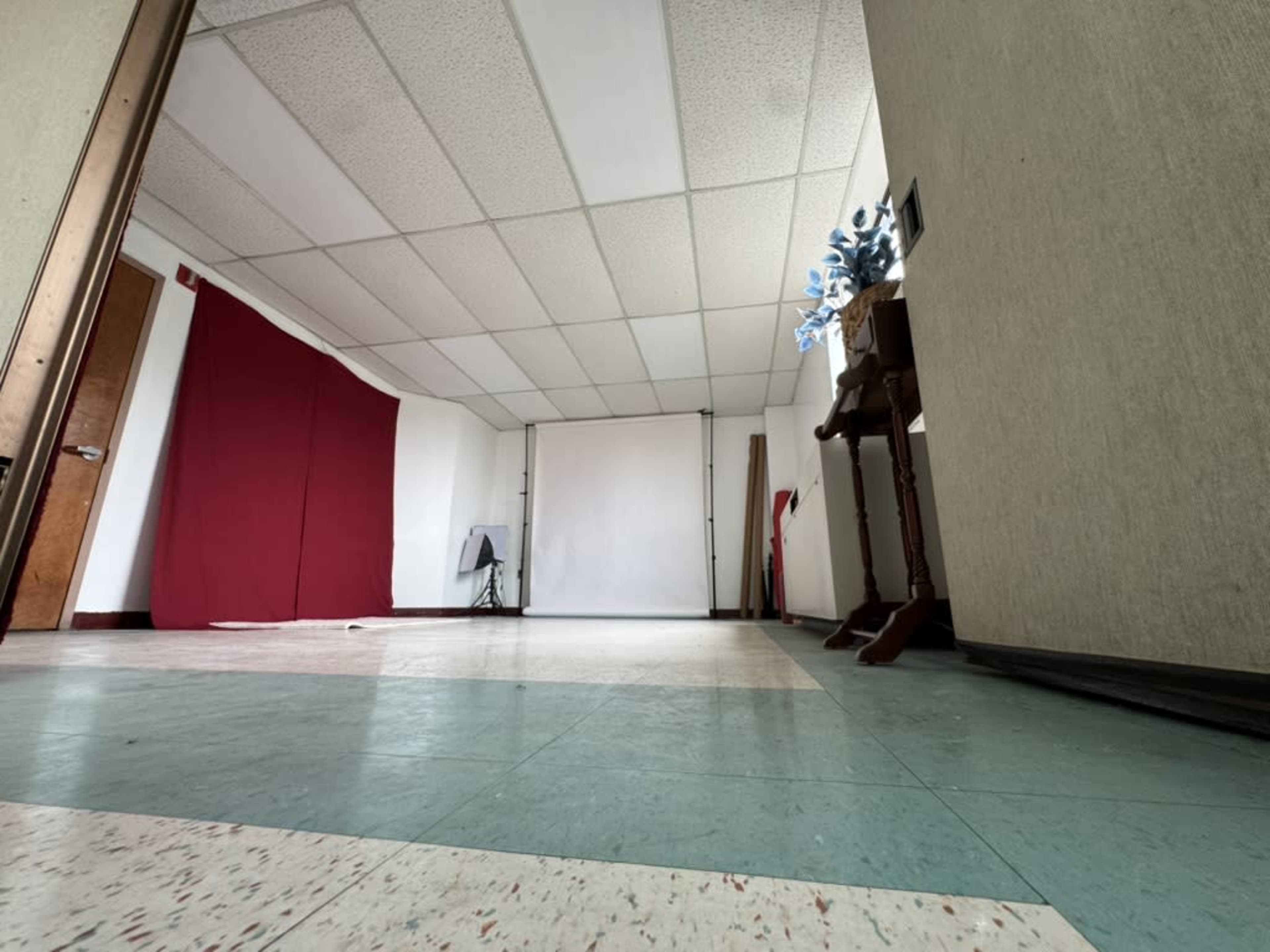 The image shows an empty room with a green and white tiled floor, a red curtain against one wall, and a backdrop setup at the far end.