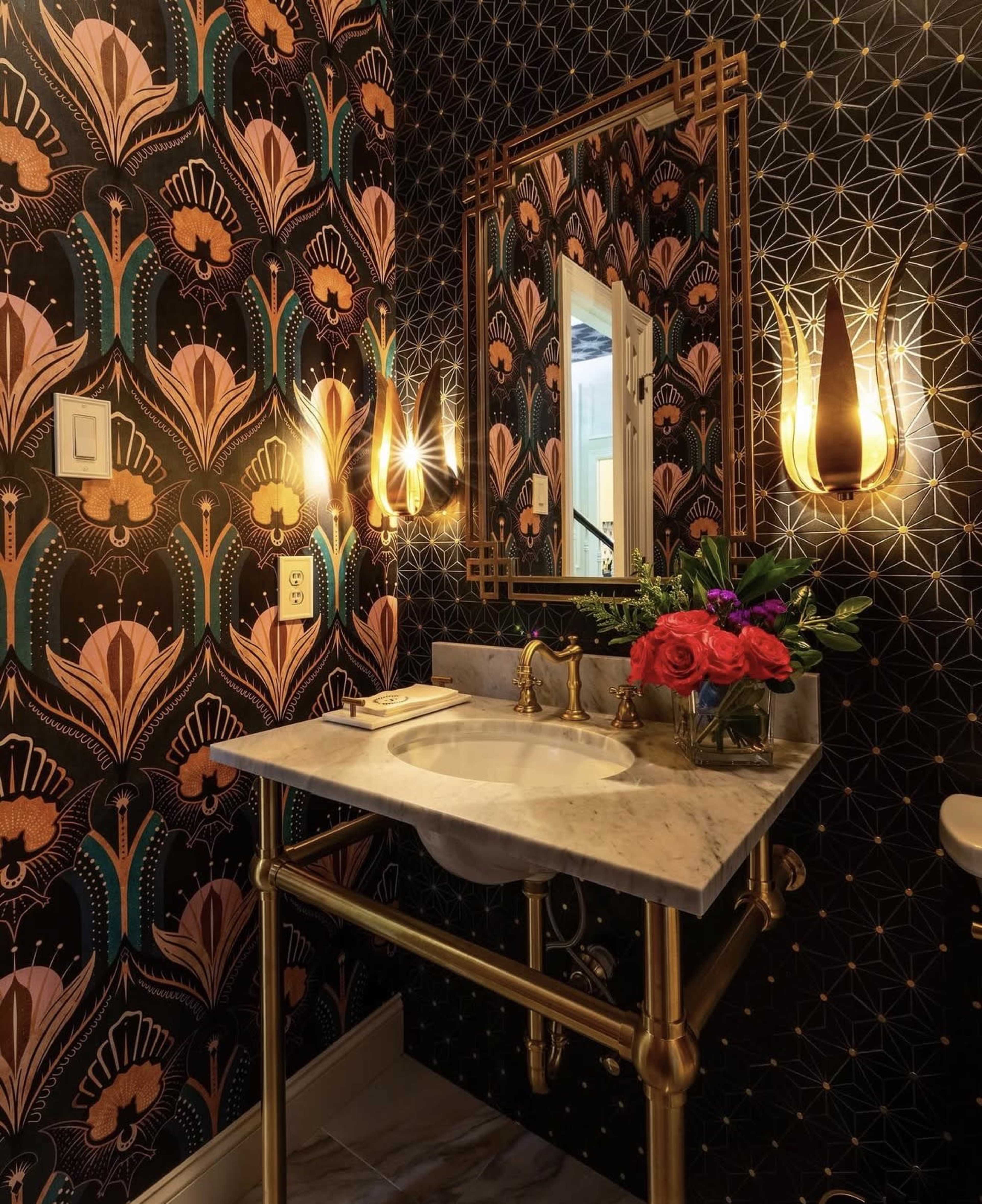 The image shows a small bathroom featuring floral-patterned wallpaper, a decorative mirror, and a marble sink on a gold frame, with two wall-mounted lights and a vase of roses on the countertop.