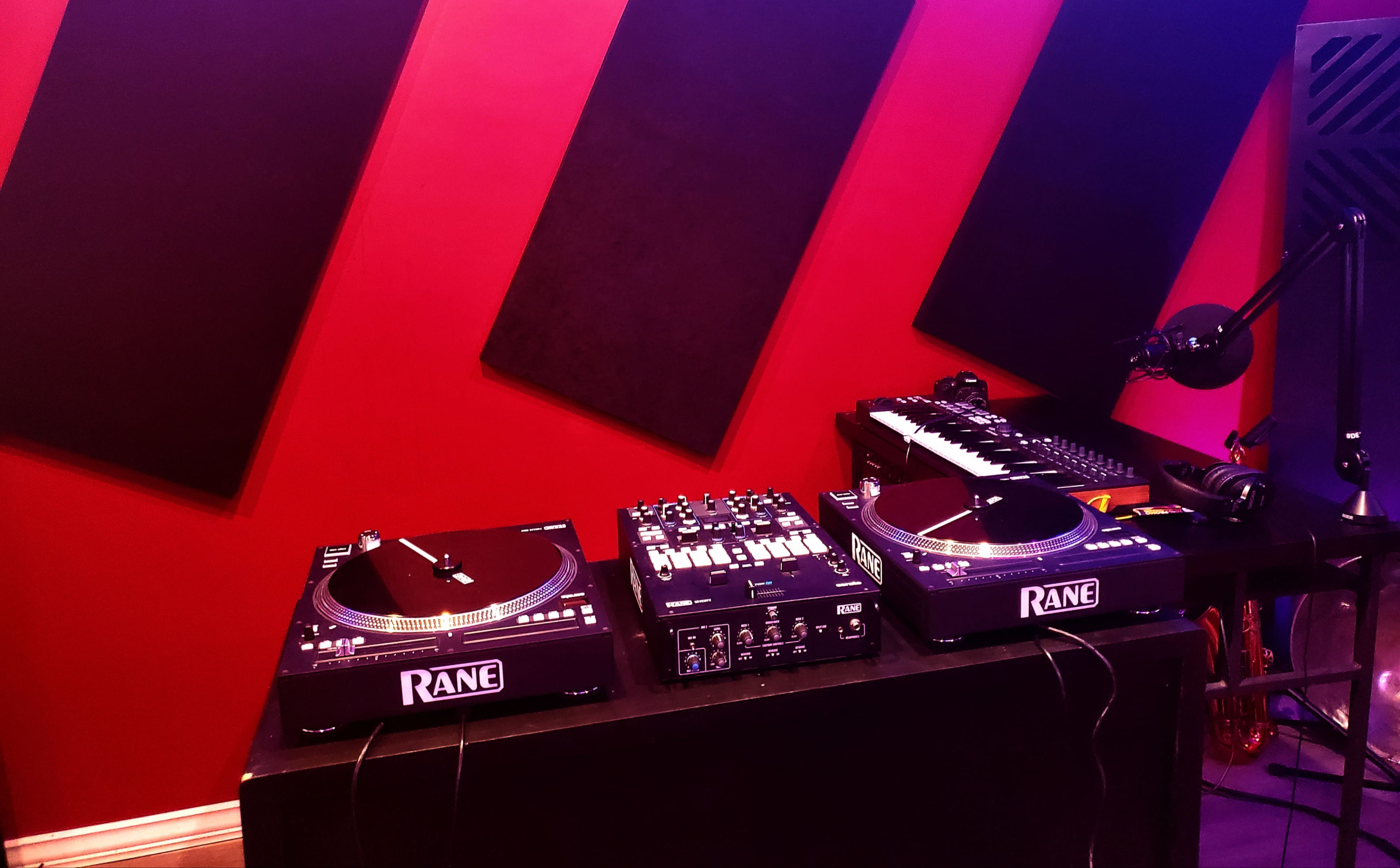 The image shows a music production setup featuring two Rane turntables, a mixer, and a keyboard against a red wall with sound-absorption panels.