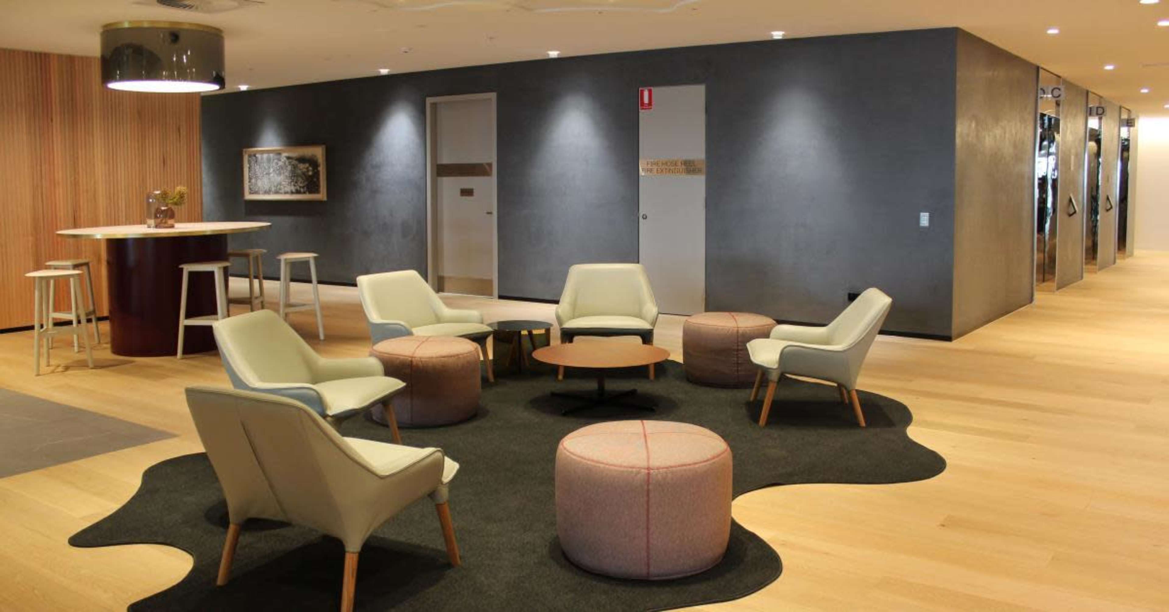The image shows a modern lounge area with several light-colored chairs, round tables, and a large area rug, surrounded by wooden accents and grey walls.