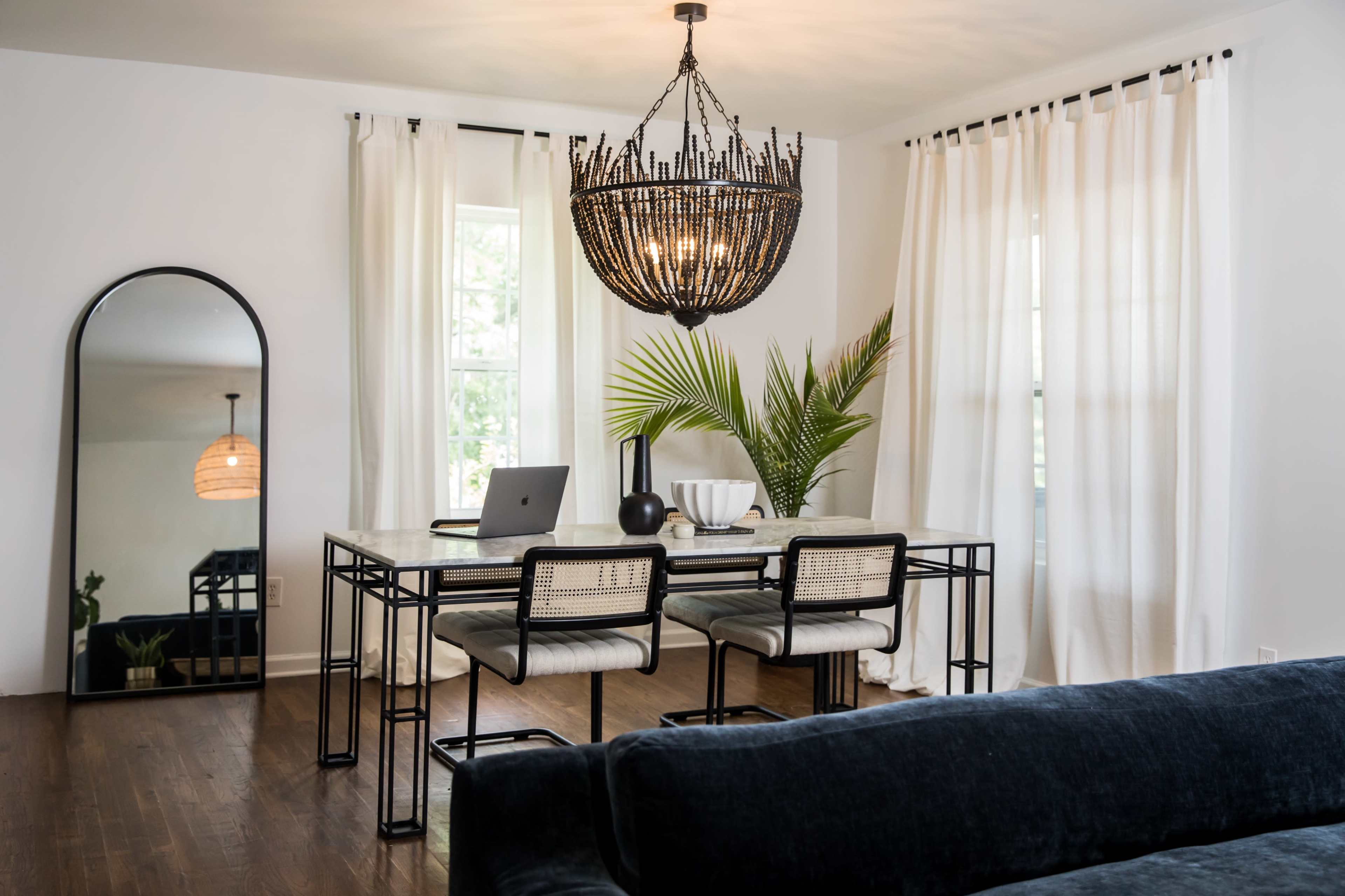 A modern dining area features a glass-top table with four chairs, a large decorative chandelier, a floor mirror, and large windows dressed with white curtains.