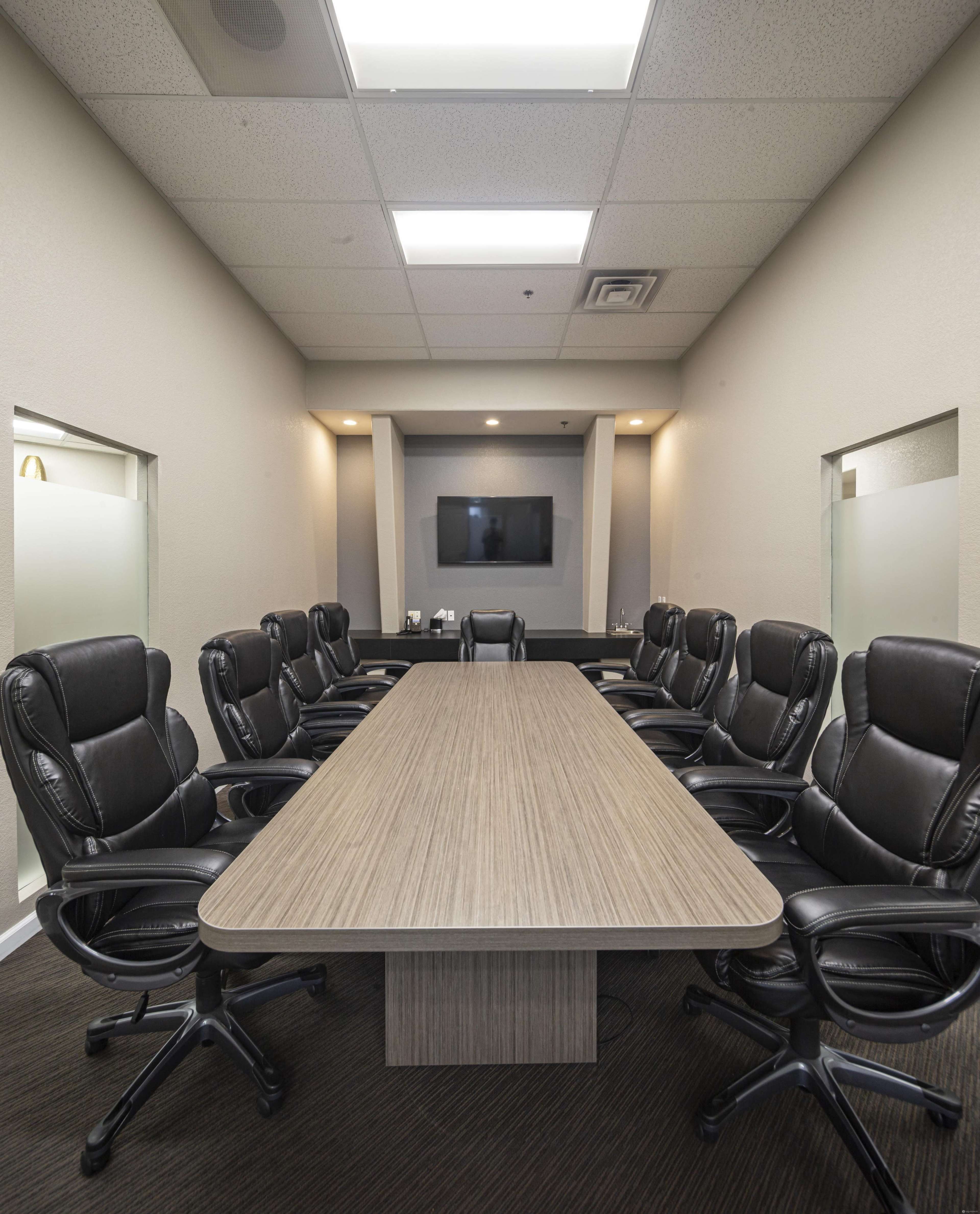 A conference room features a long wooden table surrounded by several black leather office chairs, with a television mounted on the wall at one end.