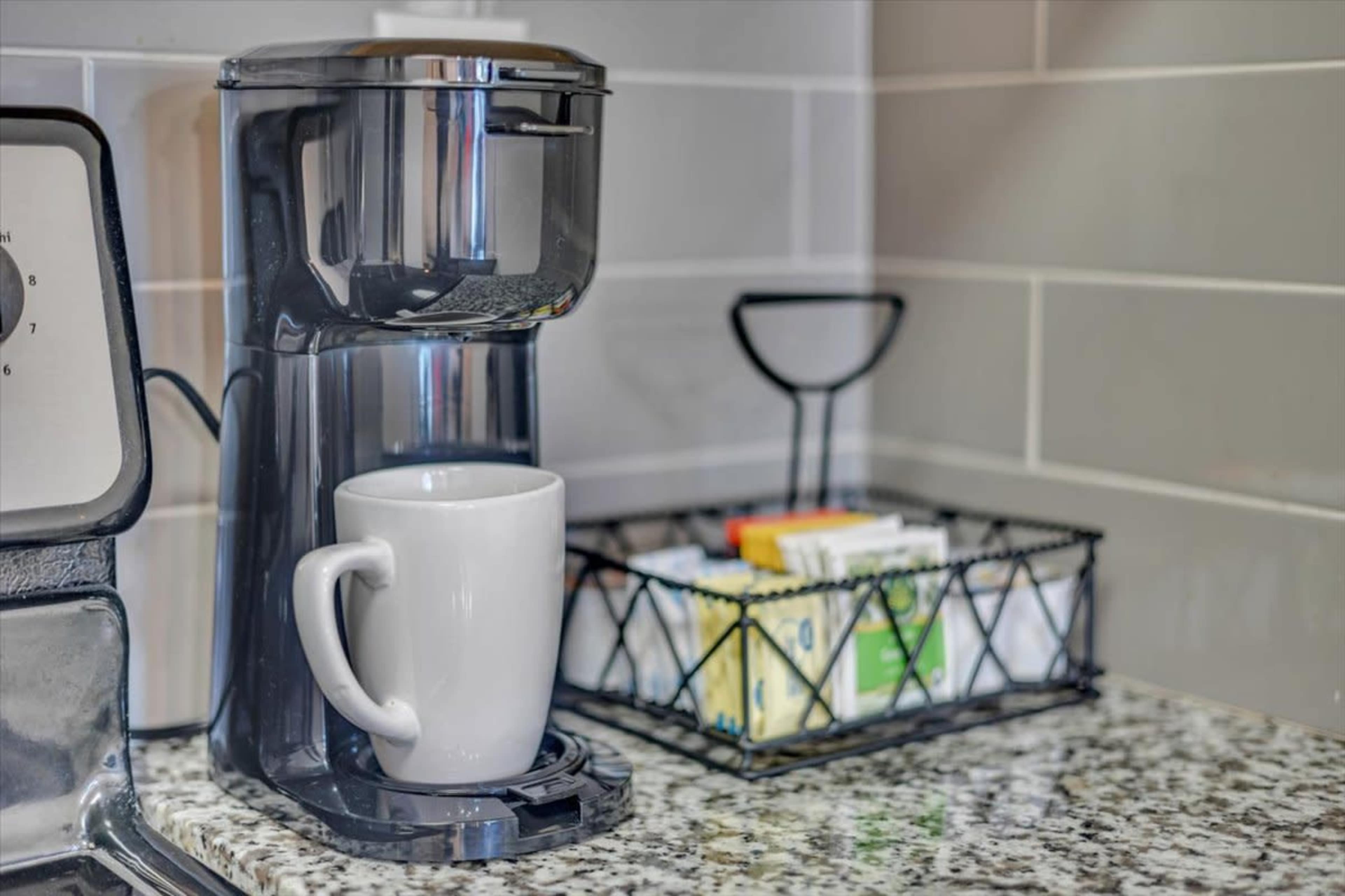 A coffee maker sits on a granite countertop next to a basket of tea bags and sweeteners.
