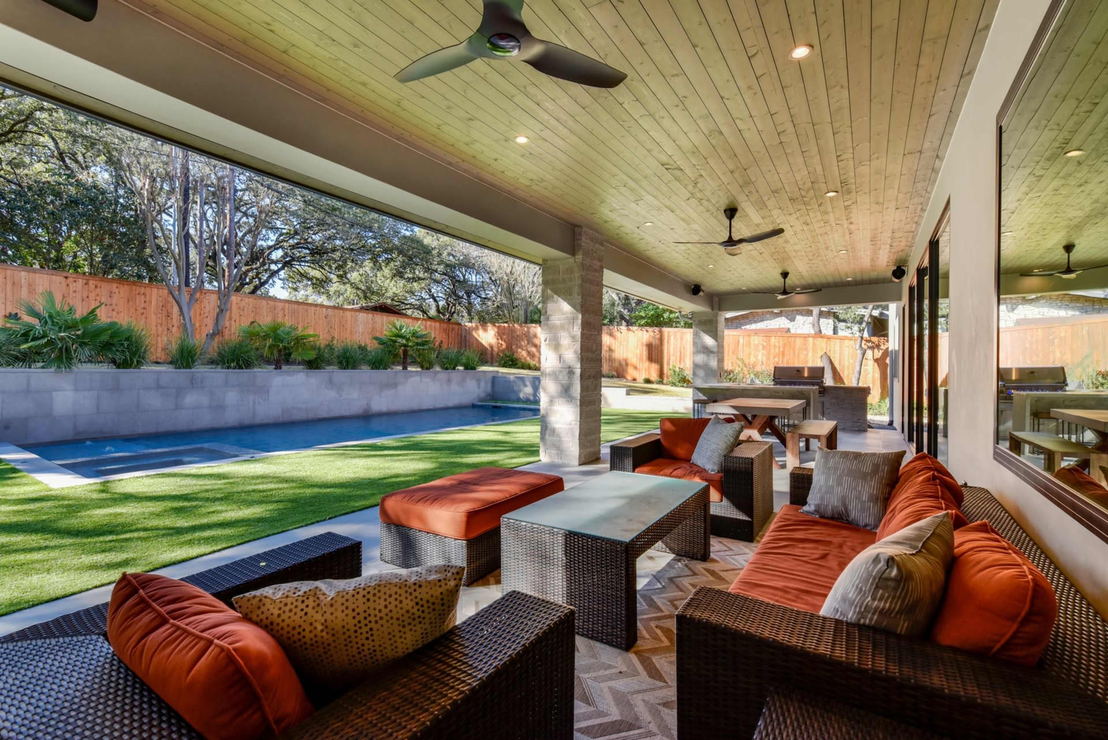 The image shows a covered patio area with seating, a coffee table, and a view of a swimming pool surrounded by landscaped greenery.