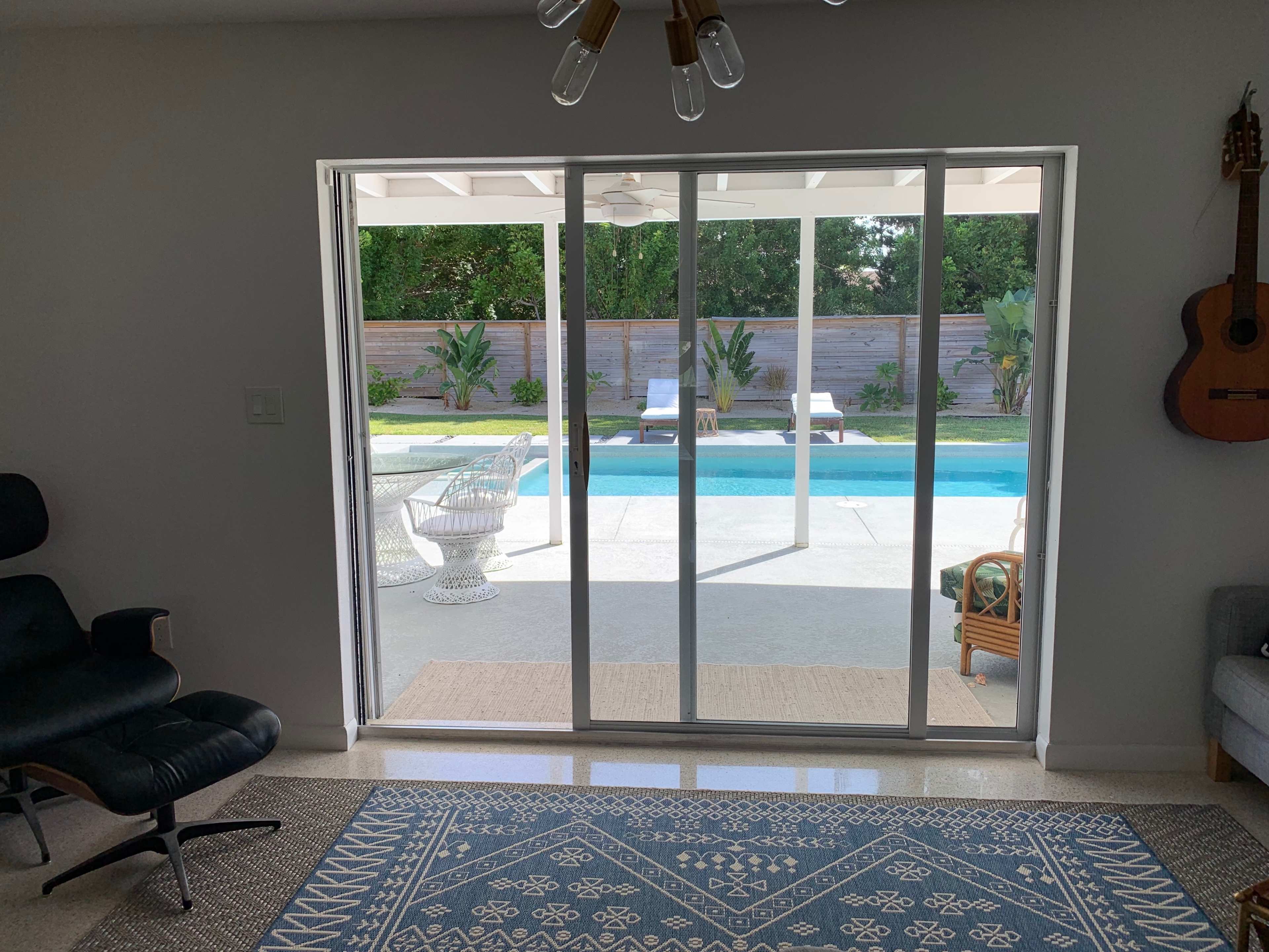 The image shows a living room with a sliding glass door that opens to a backyard featuring a pool, lounge chairs, and landscaped greenery.