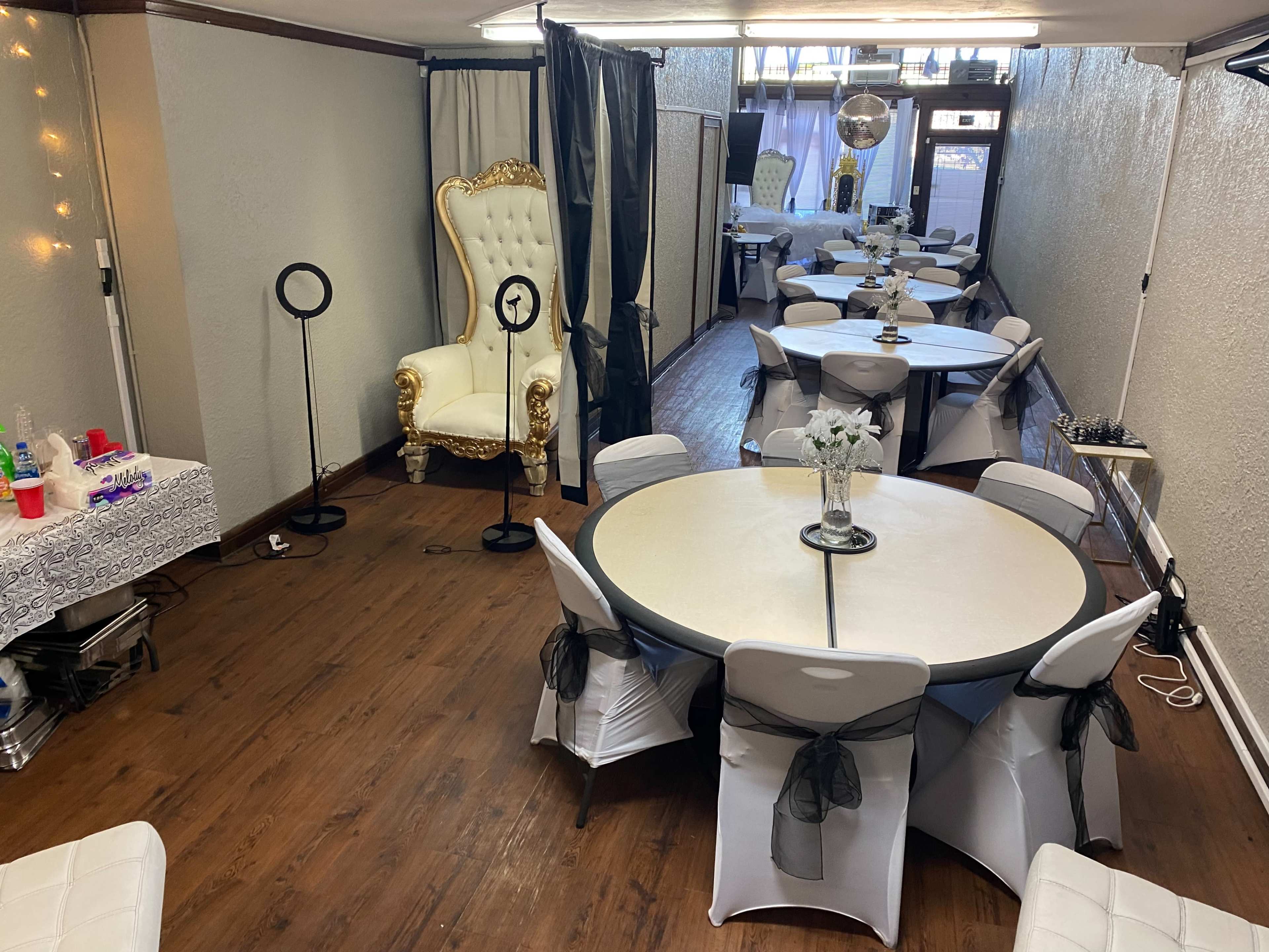 A spacious indoor event area featuring multiple round tables with white tablecloths and elegant chairs, along with a decorative throne in one corner.