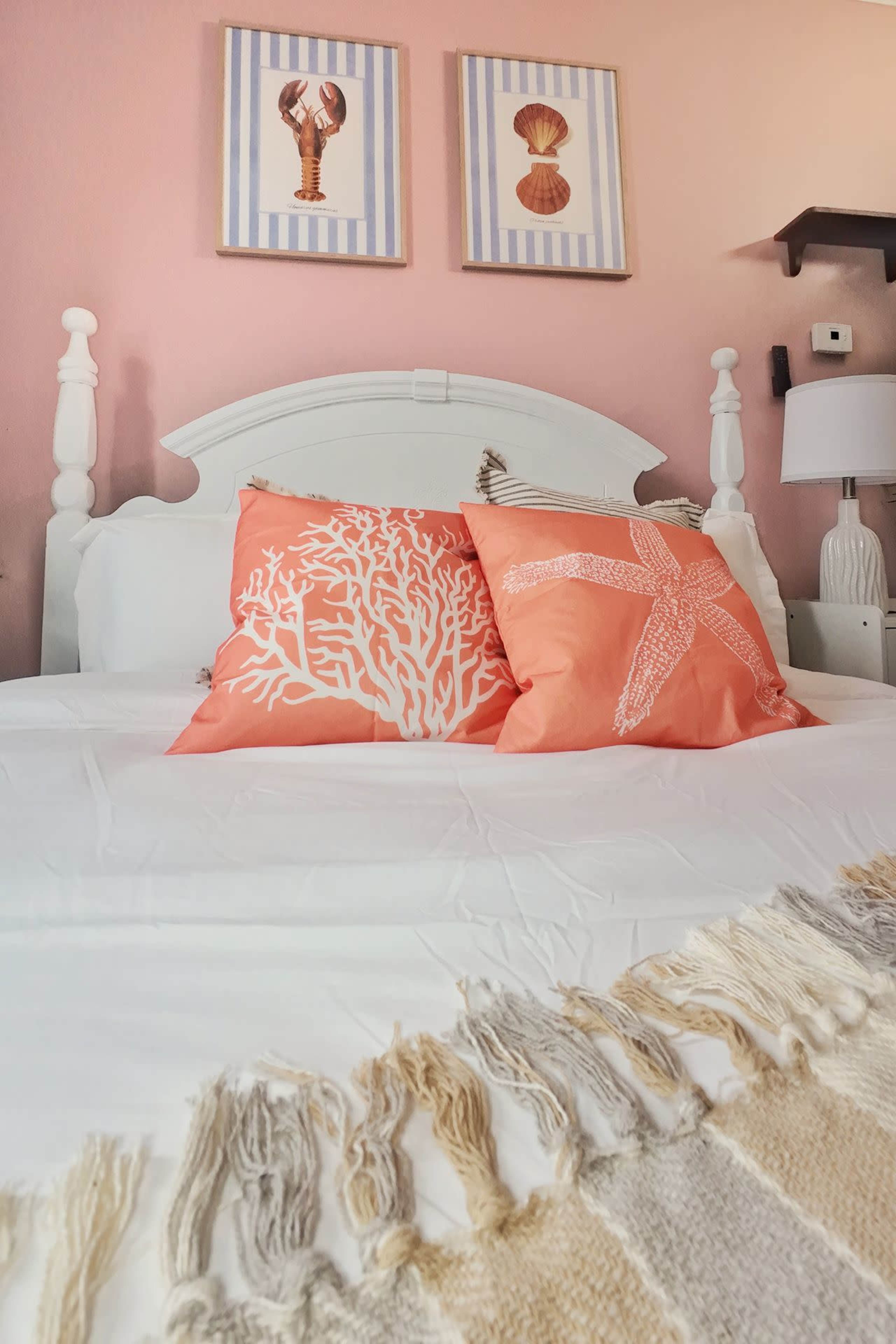 The image shows a neatly made bed with coral-patterned throw pillows and a textured blanket, set against a softly painted pink wall with framed artwork above.