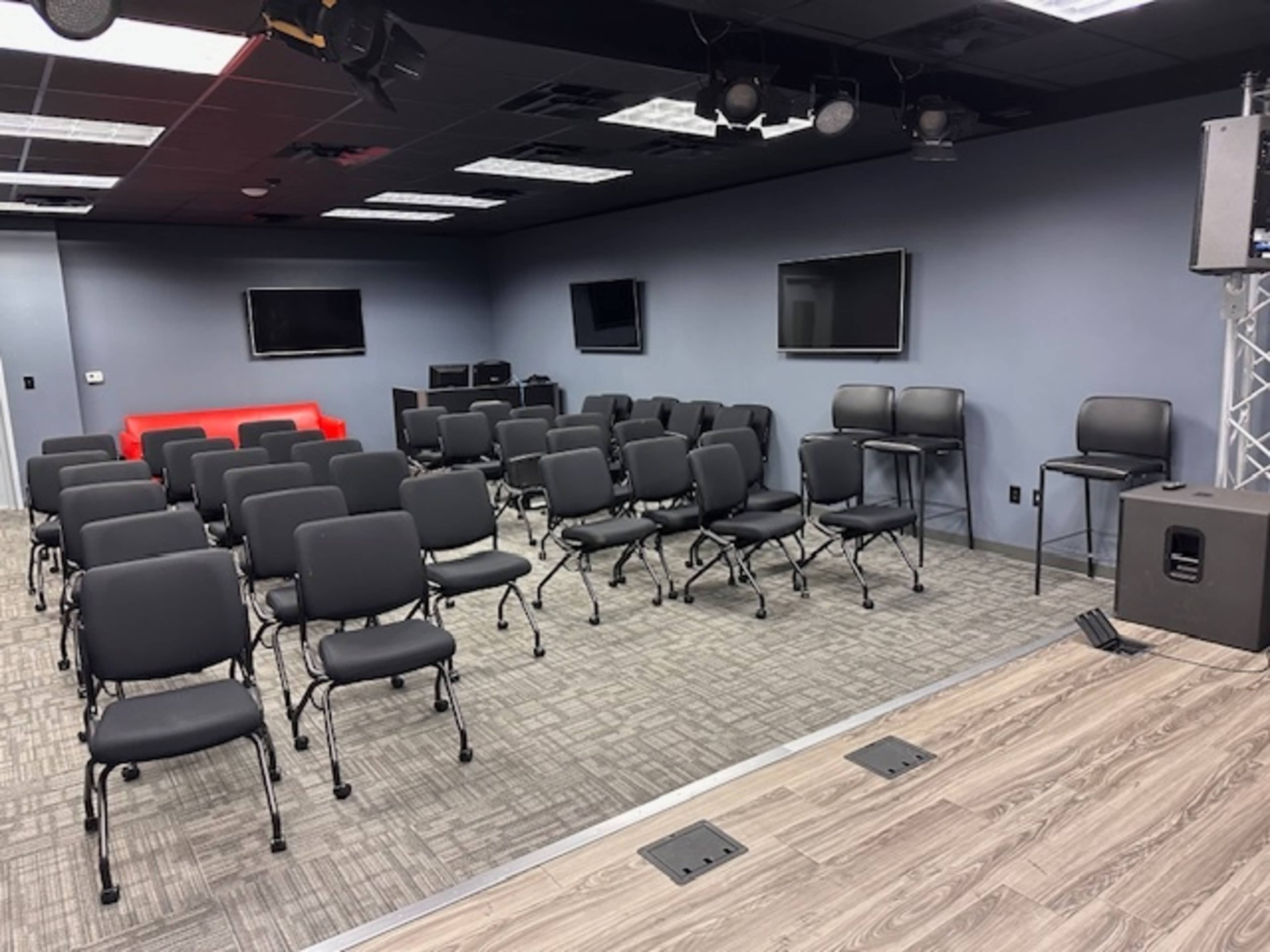 The image shows a conference room with rows of black chairs facing a small stage, flanked by two televisions on the walls.
