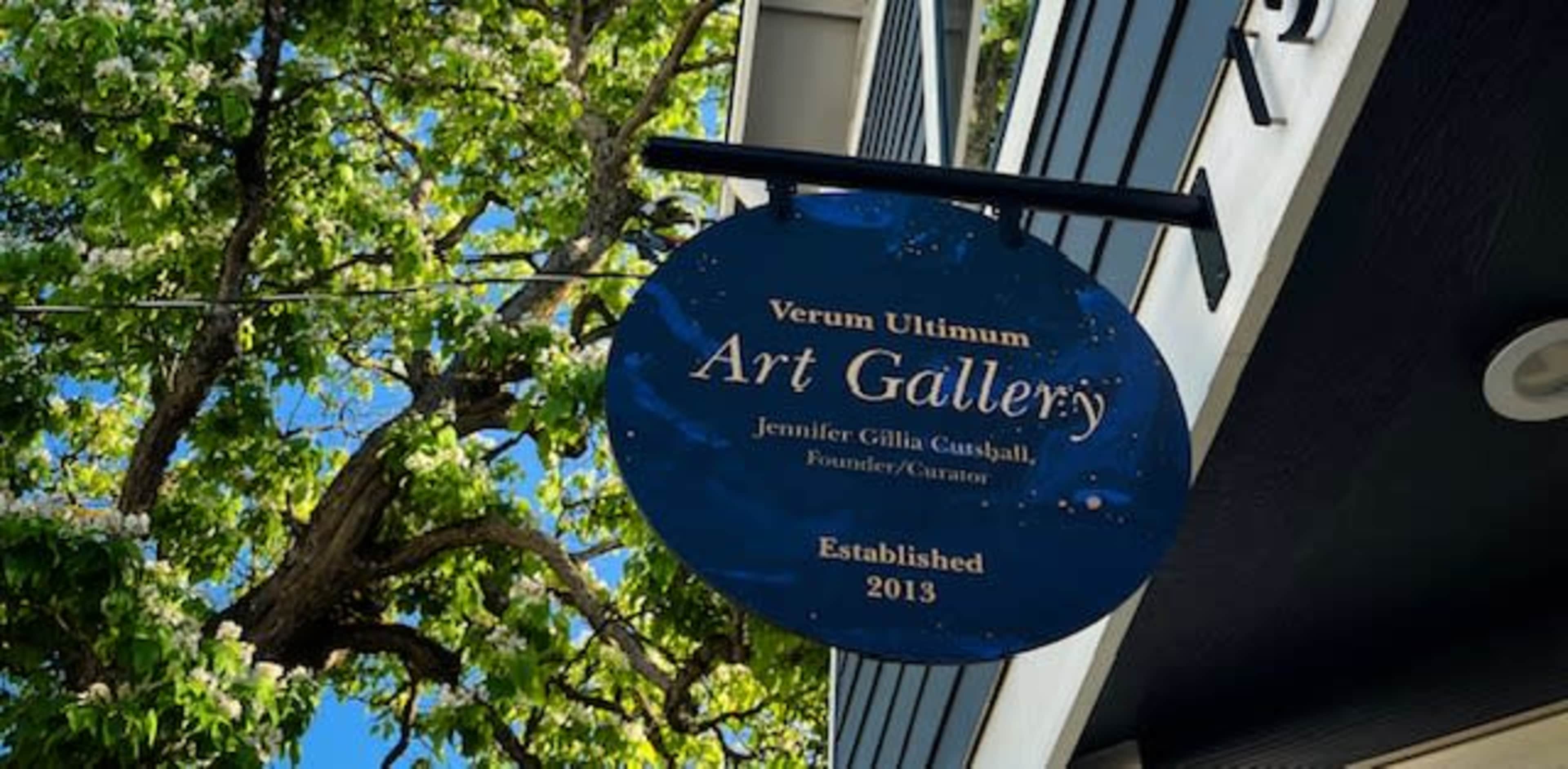 A sign for "Verum Ultimum Art Gallery" hangs in front of a building, featuring the names of the founder and curator along with the establishment year, 2013.