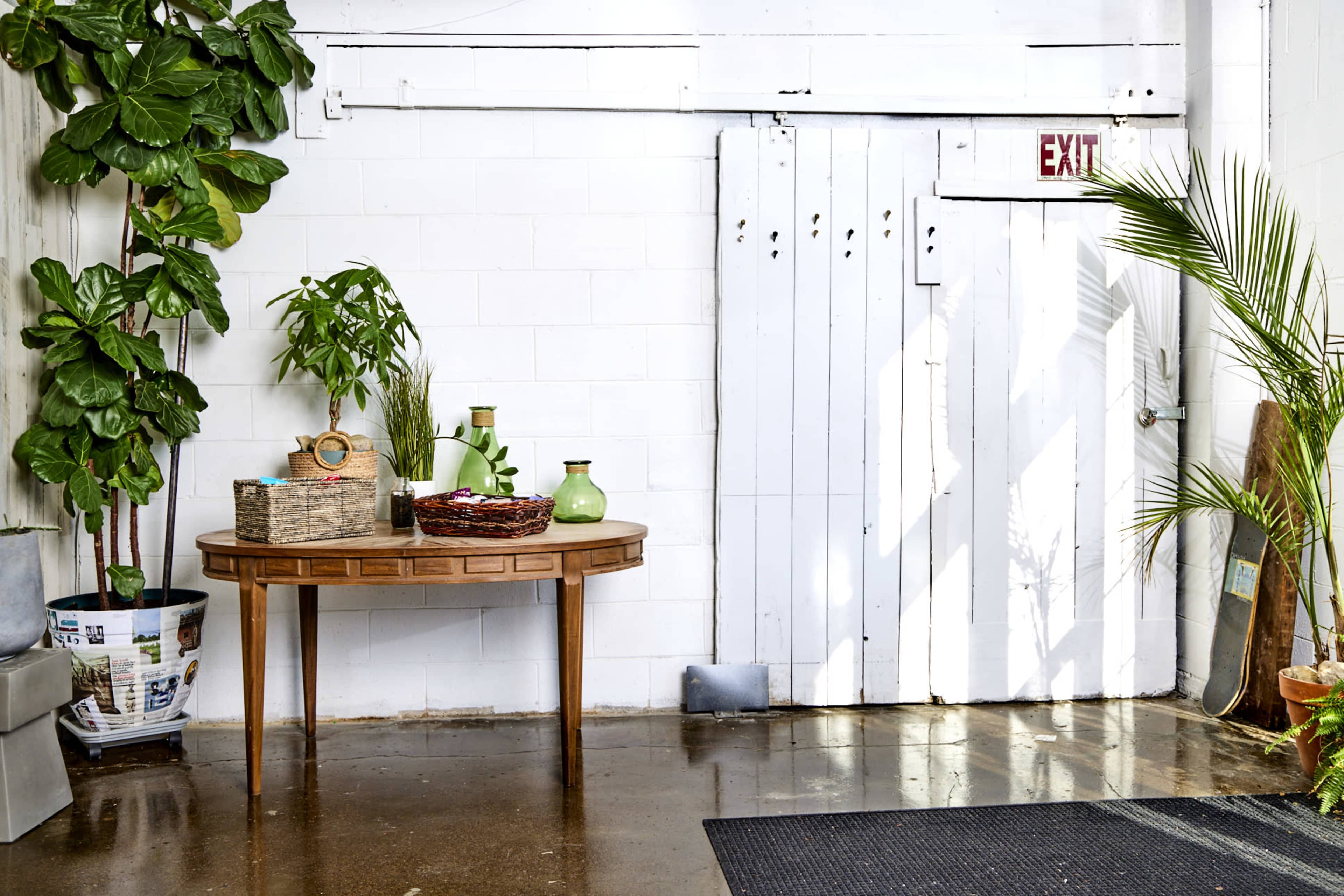 A corner of a room with a round wooden table, potted plants, and an exit door against a white brick wall.