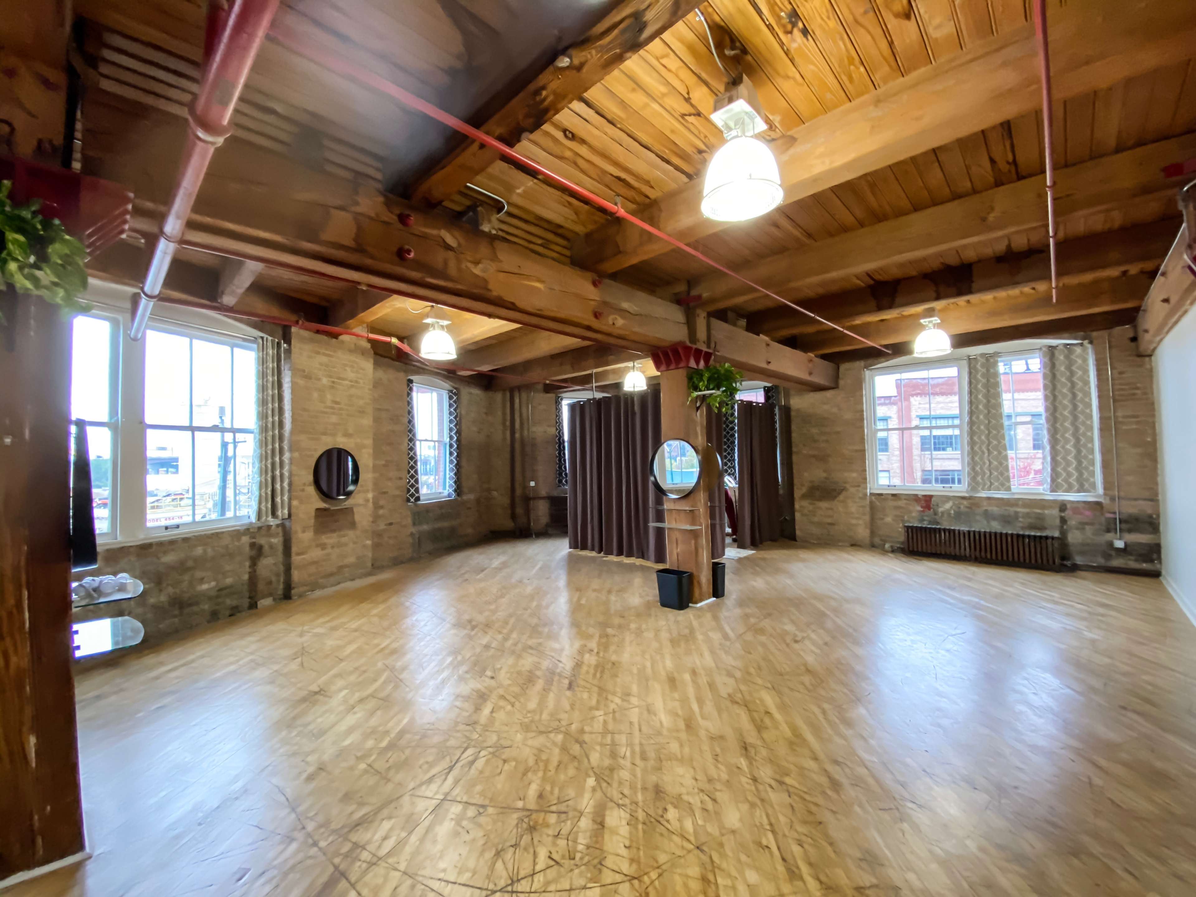 The image shows a spacious, empty room with wooden beams and floors, large windows, and industrial elements.