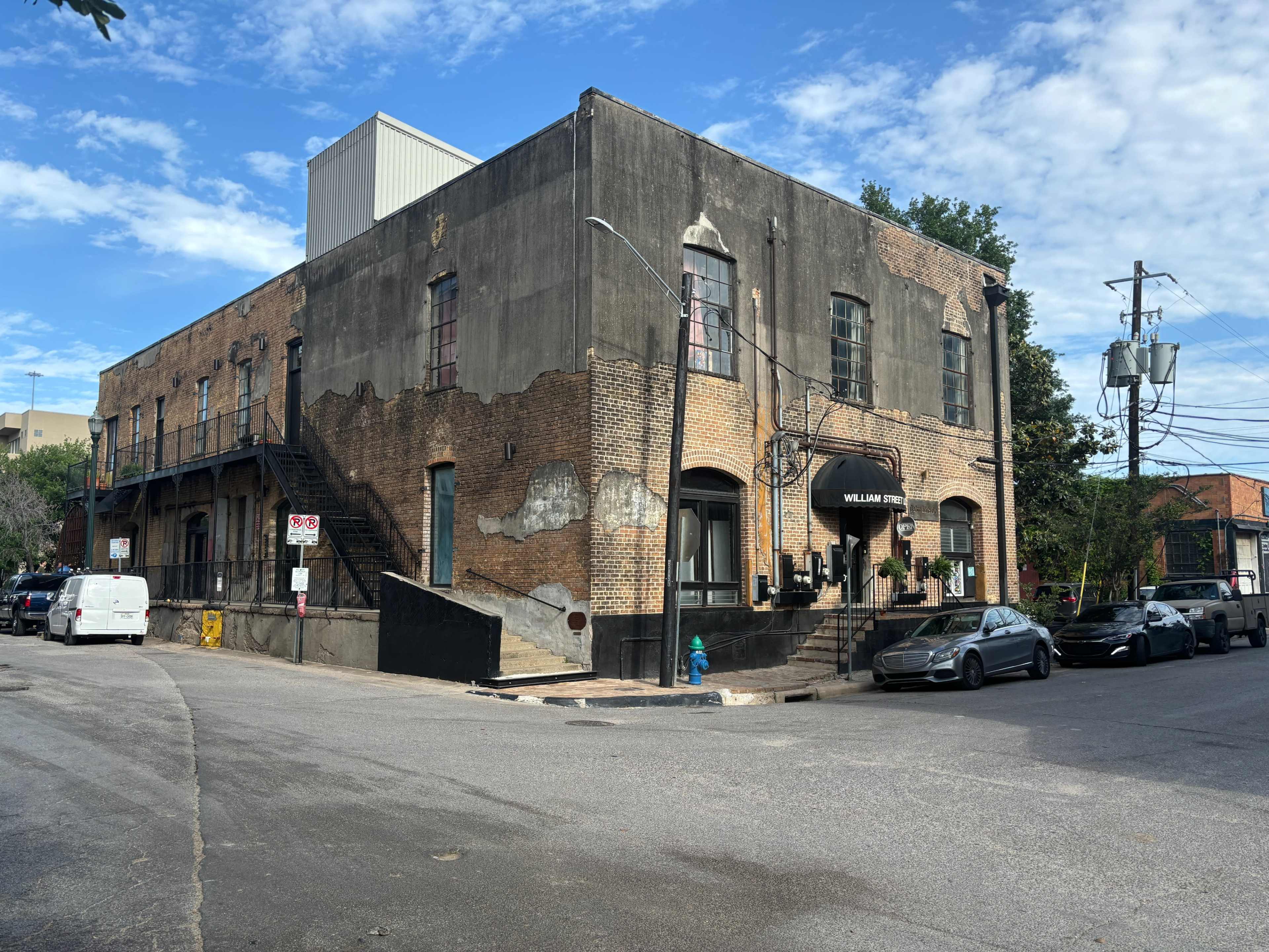 An old brick building with a weathered exterior, located at the corner of a street, featuring a staircase and a canopy entrance.