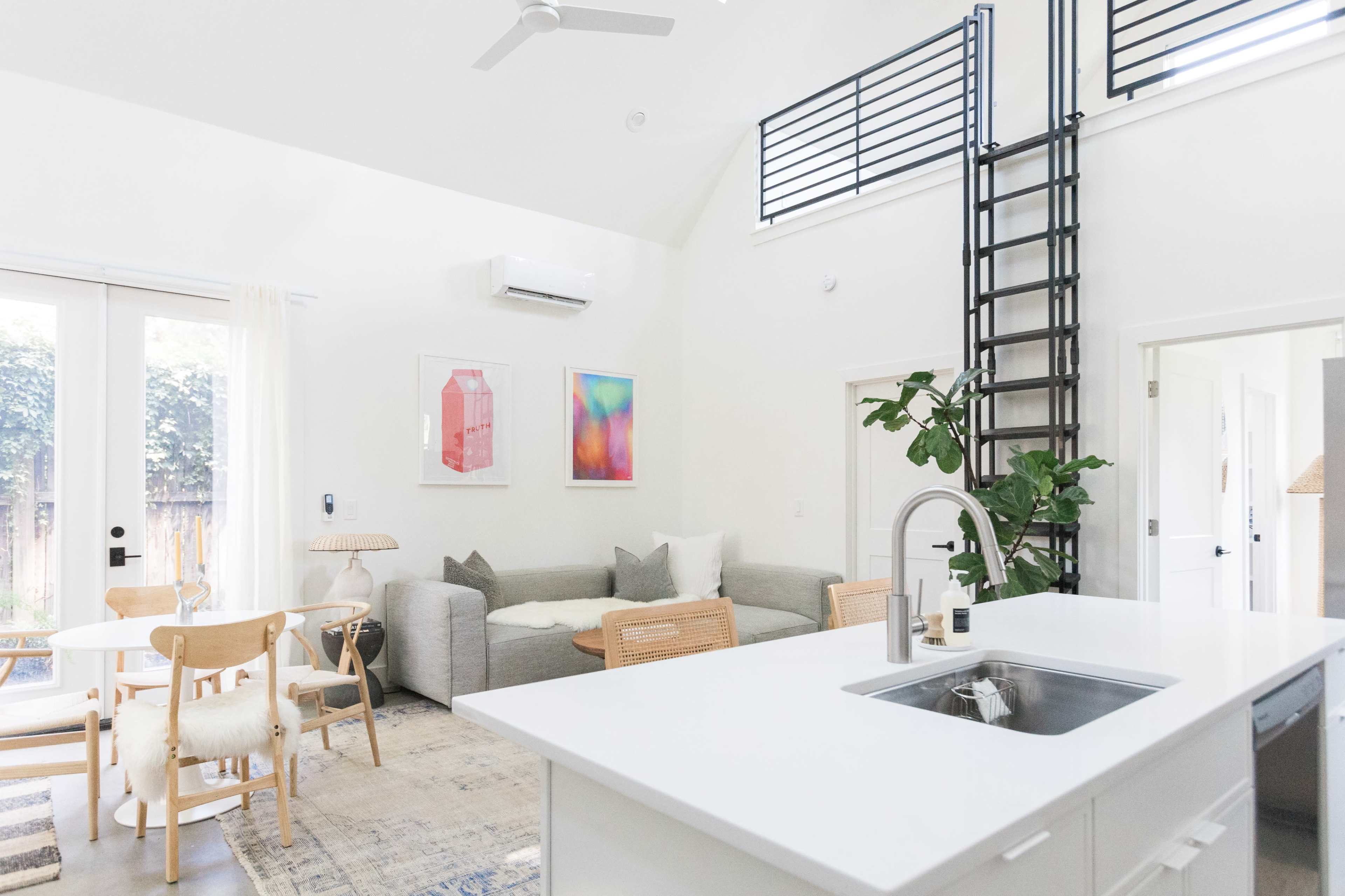 A bright, modern living space featuring a kitchen island, a dining area with light wood chairs, a cozy seating area with a gray sofa, and a ladder leading to a lofted space.