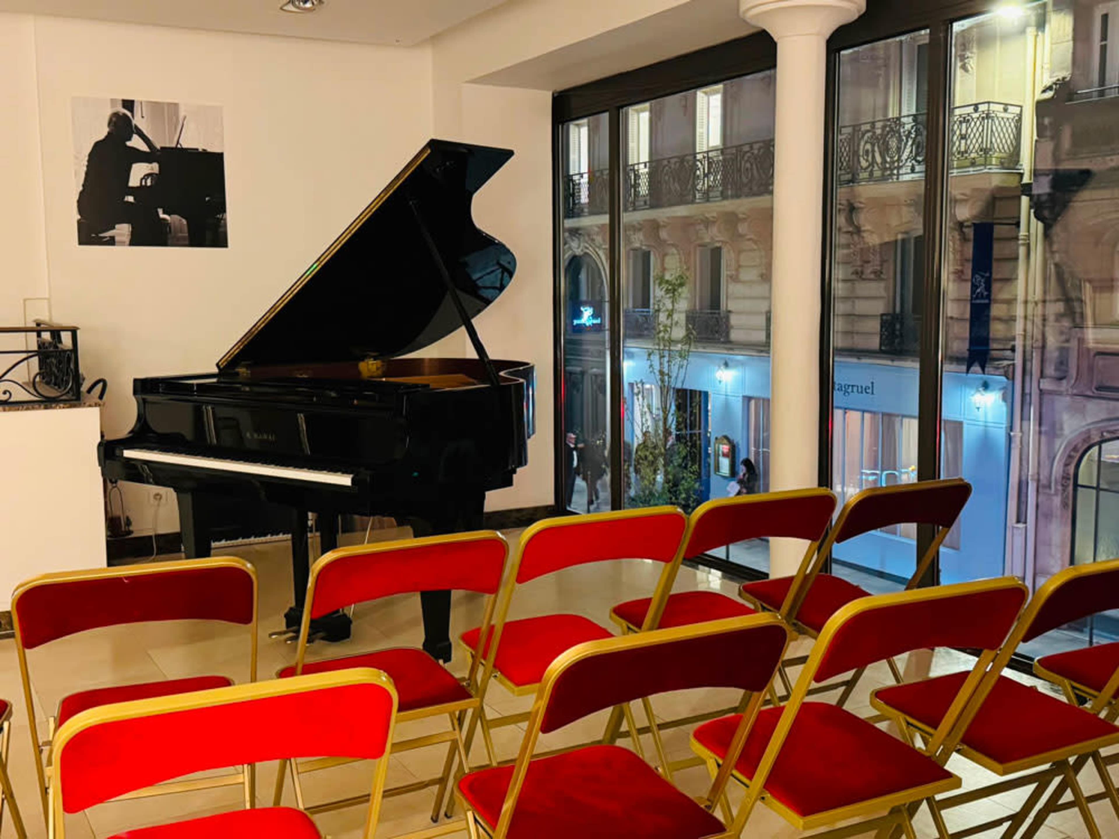 The image shows a grand piano positioned near a large window, with several red chairs arranged in front of it in a sparsely decorated room.
